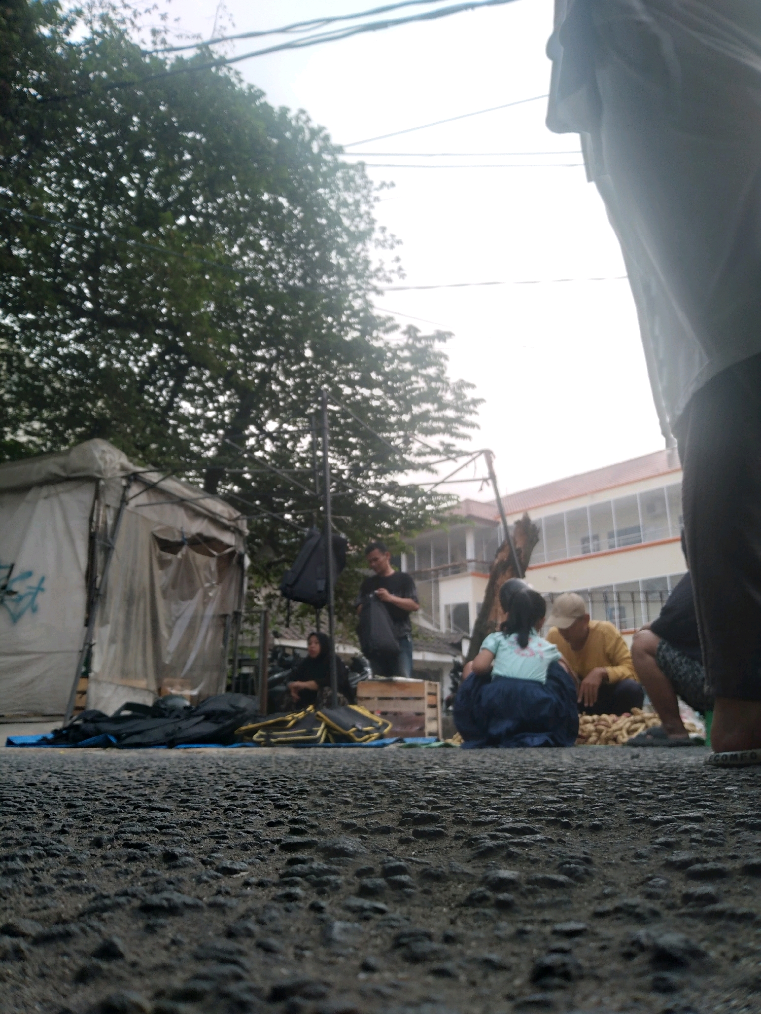 This low-angle, outdoor shot captures a street scene in South Tangerang, Indonesia, under an overcast sky. In the foreground, a rough asphalt surface dominates, suggesting a road or paved public area. Slightly further back, a blue tarp is spread on the ground, holding several black bags and a yellow folding chair.
To the left, a white tent with translucent panels and graffiti marks is set up, with wooden crates visible behind it. A metal stand with a black bag hanging from it stands near the tent.
In the mid-ground, several individuals are gathered. A woman wearing a dark hijab is seated on the ground. A man in a black t-shirt holds a black bag, looking towards the right. Further to the right, a person in a light blue top and dark blue skirt is sitting, facing away from the camera. Next to them, a man in a yellow long-sleeved shirt and a beige cap is crouching, and a person in dark patterned shorts is also seated. A pile of goods, possibly produce, is located in front of the seated individuals.
In the background, a multi-story building with white walls, balconies, and an orange roof is visible. Lush green foliage from a large tree partially obscures the building on the left side of the frame. Overhead, power lines stretch across the sky. The overall impression is a temporary market or gathering area on a street.