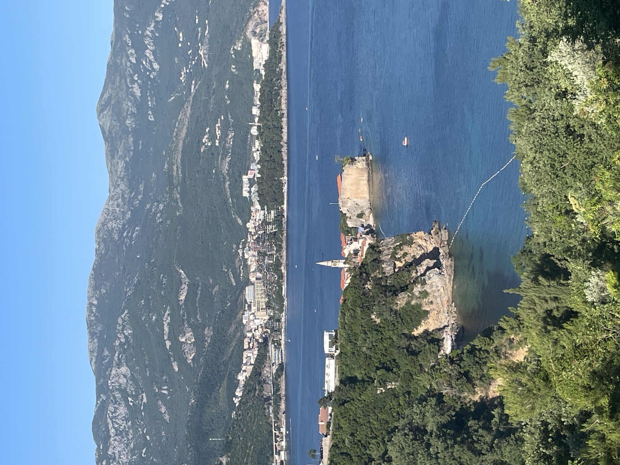 The image captures a scenic view of Sveti Stefan, a small islet and 5-star hotel resort in Montenegro, situated in the Budva municipality. The Adriatic Sea surrounds the islet. The scene includes a mountainous coastline in the background, lush green trees in the foreground, and several buildings along the coast. A line of buoys stretches out into the sea. A small boat is visible in the water near the islet. The sky is clear and blue.
