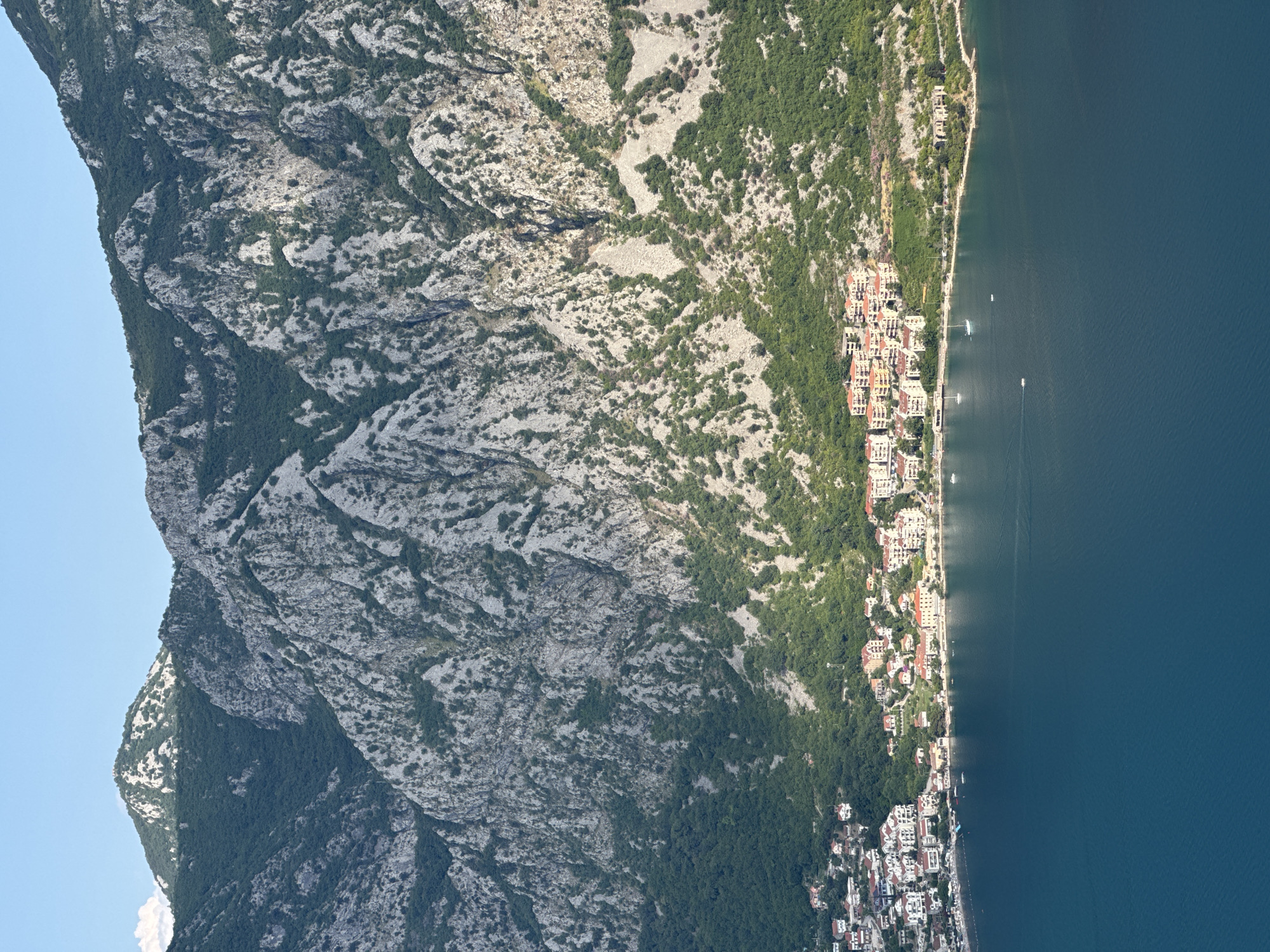 This is a high-angle, full-shot photograph of the Bay of Kotor, taken from a vantage point looking towards the town of Štrp. In the foreground, the dark blue water of the bay occupies the bottom half of the image. Along the middle ground, a coastal town is nestled at the base of a steep, rocky mountain. The town is characterized by a dense cluster of buildings with red-tiled roofs, varying in color from white to orange and yellow. The buildings are situated along the coastline, with some extending further inland. A road runs along the edge of the town. Beyond the town, the imposing mountain rises sharply, its slopes covered in patches of green vegetation and interspersed with barren, grey rock faces. The sky above is a clear, pale blue. The scene depicts a peaceful daytime view of a coastal settlement set against a dramatic mountainous backdrop.