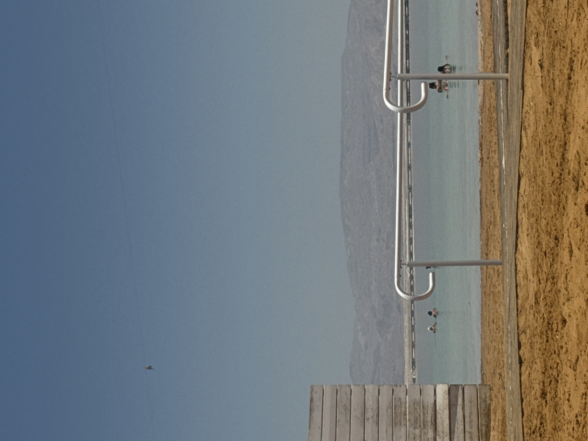 The image shows a beach scene at the Dead Sea in Tamar Regional Council, Israel. The foreground features a wooden boardwalk leading to the sea, with a metal handrail visible. To the right is a sandy beach. The Dead Sea occupies the middle ground, with several people floating in the water. The background shows mountains or hills on the opposite shore under a clear, blue sky. A single bird is visible in the sky.
