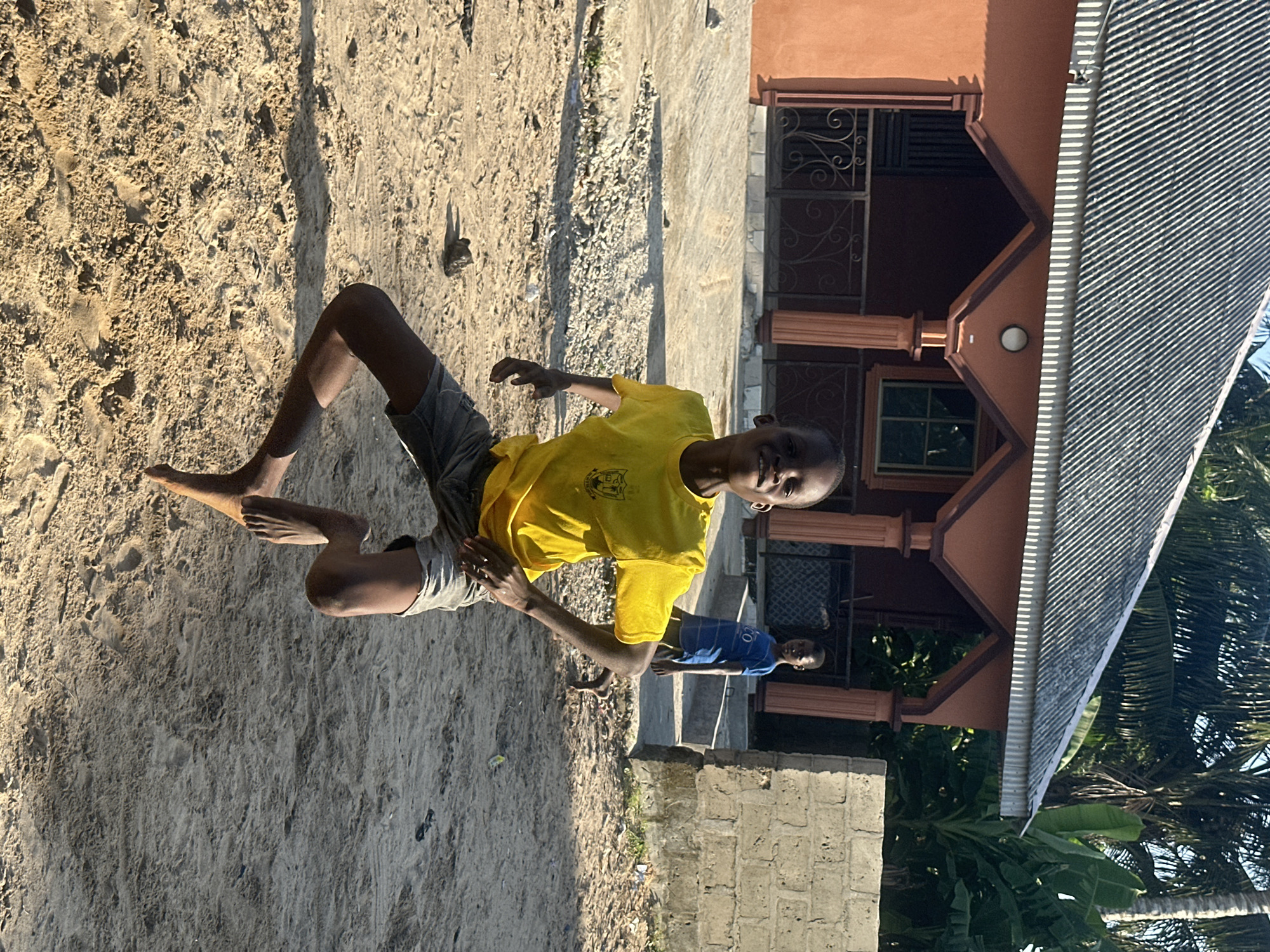 The image features two young boys outdoors in what appears to be a sandy, possibly beach-like area, with a building in the background. The primary subject is a boy in the foreground, wearing a bright yellow t-shirt and grey shorts. He is captured in mid-action, with his body angled and one leg extended, suggesting he is playing or jumping. His expression is unclear due to the angle and lighting, but he appears to be looking towards the camera. The sandy ground is textured with footprints and small debris.
Behind the main subject, a second boy is visible, wearing a blue t-shirt and shorts, standing further back. He is also looking in the direction of the camera.
The building in the background is a multi-story structure with an orange-brown facade and a metal roof. It has decorative elements, including patterned window grills and pillars. Lush green vegetation, including palm trees, is visible around the building, indicating a tropical or subtropical environment.
The lighting suggests it is daytime, likely late afternoon, given the long shadows cast on the sand. The weather appears to be sunny and warm. The scene evokes a sense of playful activity in a relaxed, outdoor setting, possibly a residential area near the coast in Badagry, Nigeria. No visible text is present in the image.