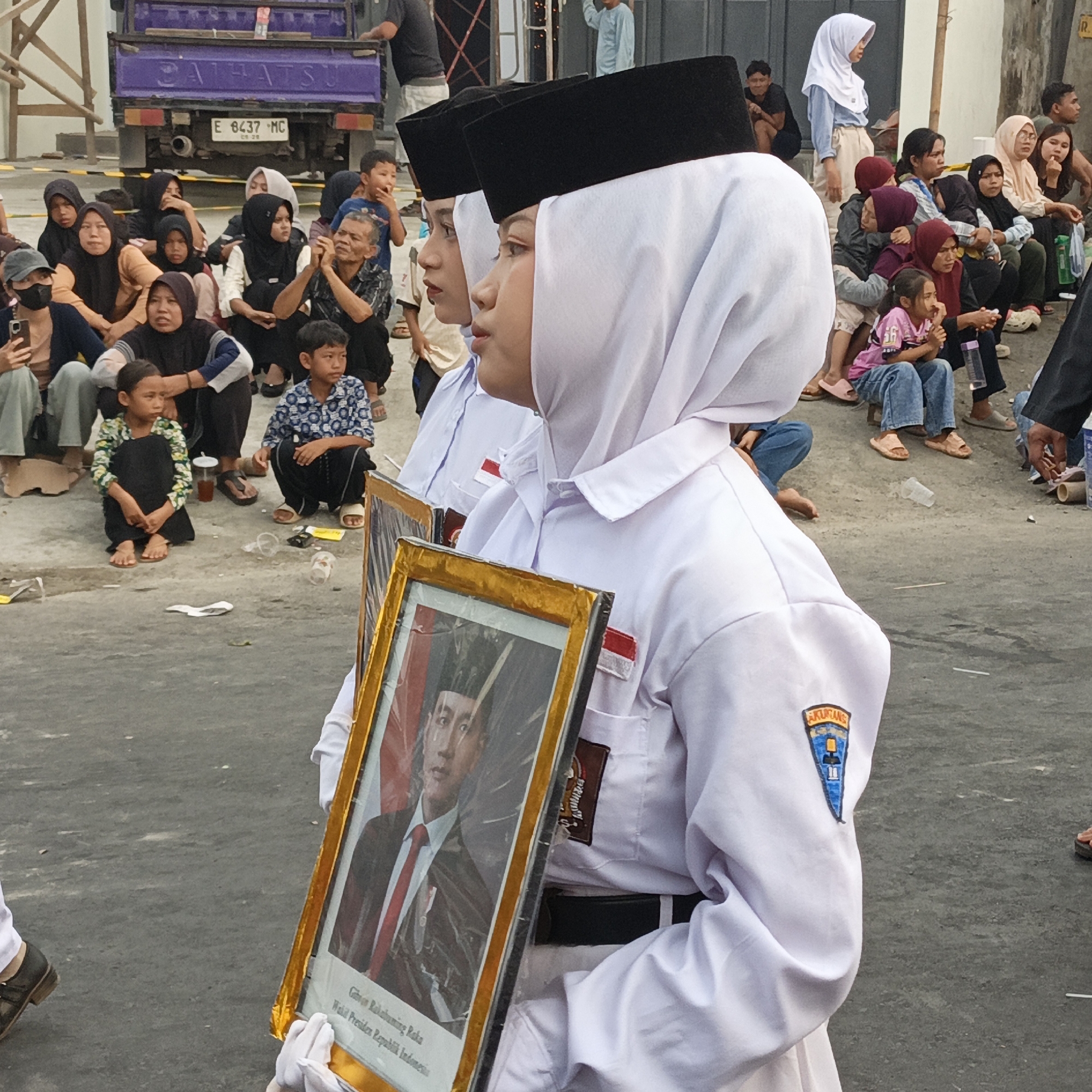 The image captures a group of people gathered outdoors in what appears to be an urban or semi-urban setting, possibly in Randudongkal, Indonesia. In the foreground, two individuals in white uniforms and black skullcaps, likely students, are prominently featured. One of them is holding a framed portrait of a man in a suit and tie. The portrait is partially visible, showing the man's face and a red tie. The background reveals a purple truck with license plate "E 6437 MC" and some onlookers seated on the ground, many of whom are wearing headscarves. The ground is paved and littered with some debris, including plastic cups. The overall atmosphere seems to be that of a public gathering or event.