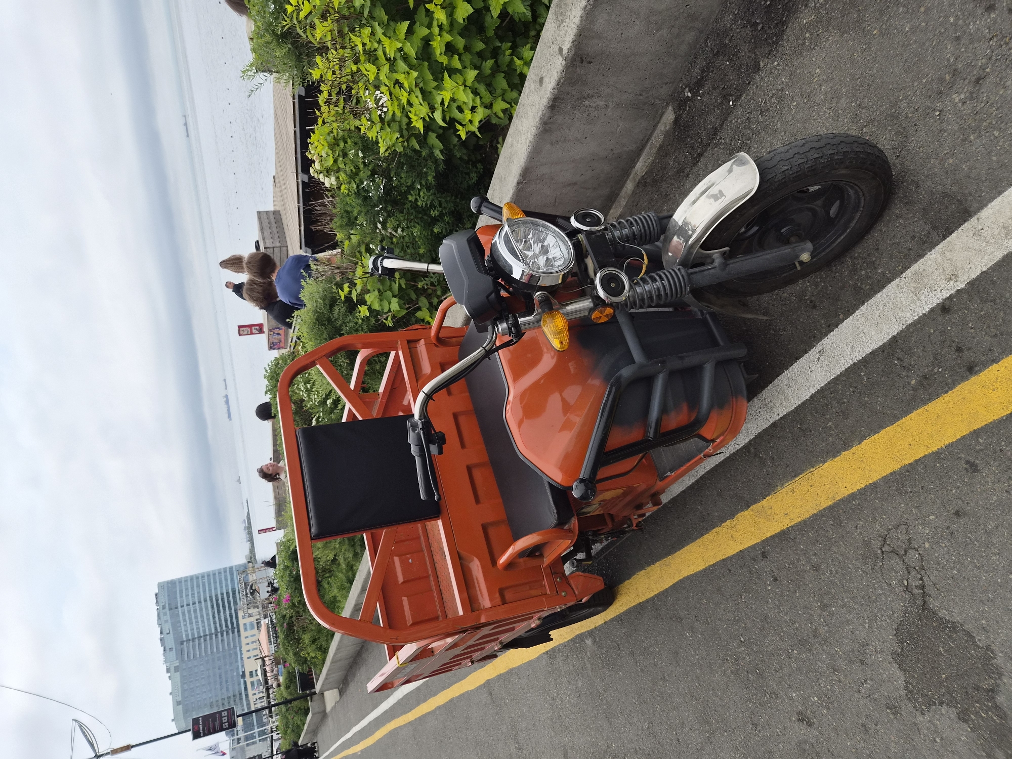 In Vladivostok, Russia, a three-wheeled orange cargo scooter with black seating is parked on an asphalt road marked with yellow and white lines. The scooter has a metal frame around its cargo bed and a single headlight. In the background, a modern, glass-facade building overlooks a body of water with several ships visible on the horizon. A wooden deck extends into the water, with a few individuals standing and sitting on it. Lush green bushes line the edge of the road and the deck. The sky is overcast.