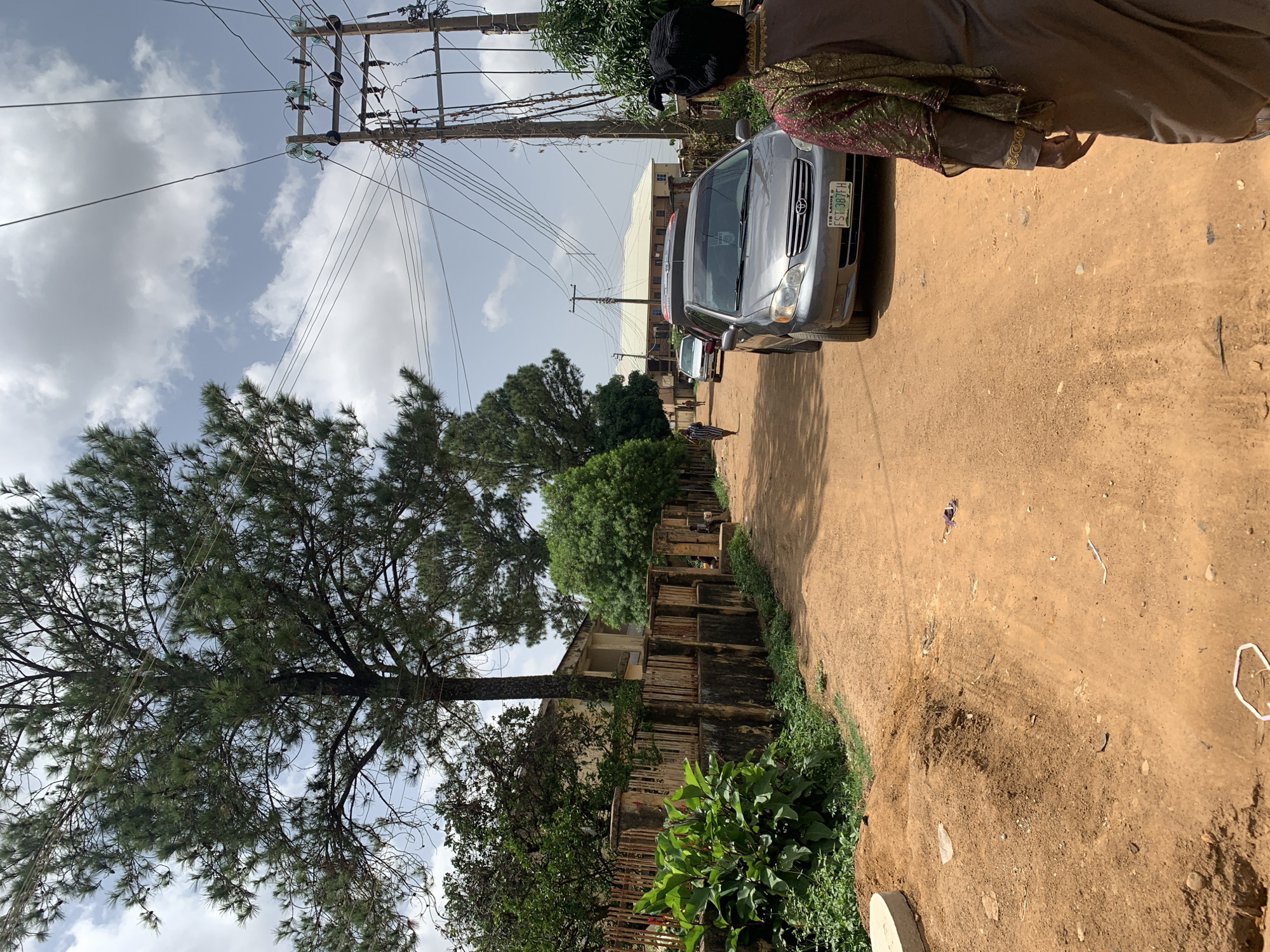 The image shows a dirt road in Rafin Par, Nigeria, with a parked silver Toyota Corolla sedan and other vehicles visible in the distance. A person wearing traditional clothing is partially visible in the top right corner of the frame. Utility poles with power lines run alongside the road, and trees and foliage are present on the left side. A building with a deteriorating fence is also visible along the road. The sky is partly cloudy.