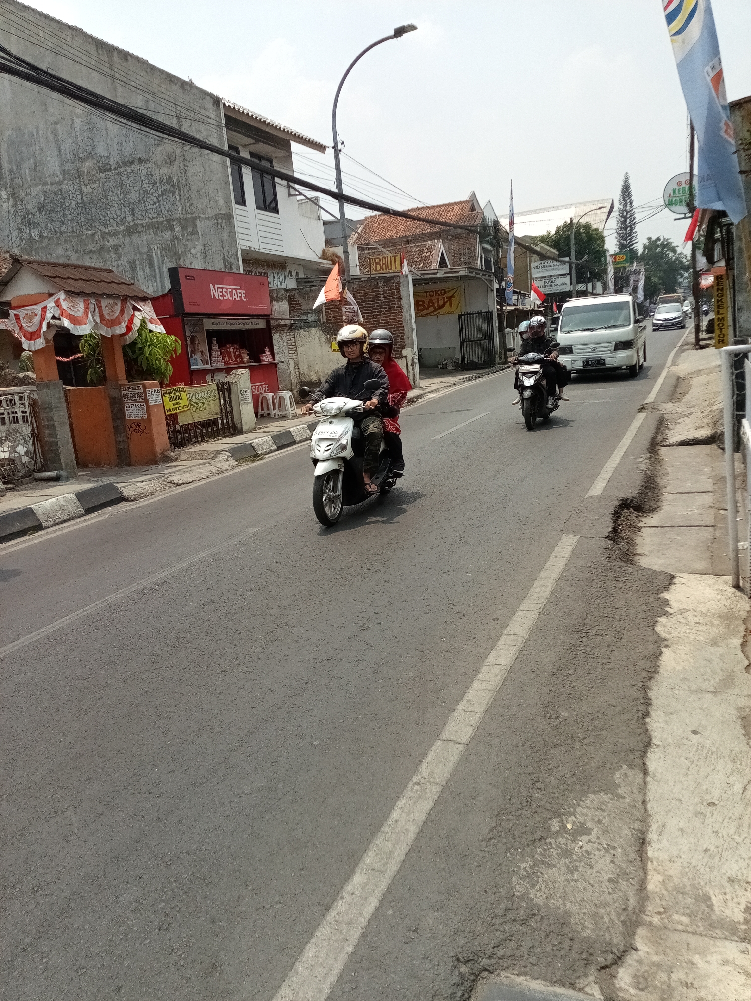 The image depicts a street scene in Cimahi, Indonesia, on a bright, overcast day. The foreground is dominated by a two-lane asphalt road. On the left side of the road, a white scooter is being ridden by a person wearing a yellow helmet and a dark jacket, with another person in a red shirt behind them. The scooter has a license plate that appears to read "D 8852 SGD".
Adjacent to the road on the left is a sidewalk and a row of buildings. One building features a red Nescafé stall with various products displayed. Another building has a sign that reads "TOKO BAUT". Further down the street, there are other shops and businesses.
On the right side of the road, a white van is traveling away from the camera. The van has a license plate that reads "D 8753 UF". Other vehicles, including cars and motorcycles, are also visible further down the road. The environment appears to be a commercial and residential area with trees and utility poles visible in the background. The overall impression is of a busy urban street with moderate traffic.