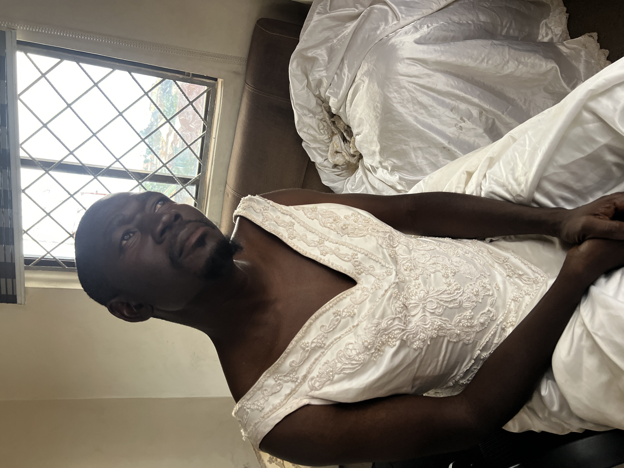 A dark-skinned man is sitting down, facing the camera, wearing a white wedding dress. He has a shaved head and is looking to his left, with his chin tilted upwards. He has a light beard. The dress is sleeveless and has a V-neckline, decorated with lace and pearls. The man's arms are resting on his lap. To his right, on what appears to be a bed, is a large pile of white fabric, possibly part of another dress or bedding. Behind the man is a window with a diamond-patterned grille, showing a view of a city with buildings and vegetation under a bright sky. To the left of the man is a beige wall. The lighting in the room is dim, with light coming from the window. The overall impression is that the photo was taken indoors, in a residential setting within the city of Jos, Nigeria, as indicated by the architecture and vegetation visible through the window.