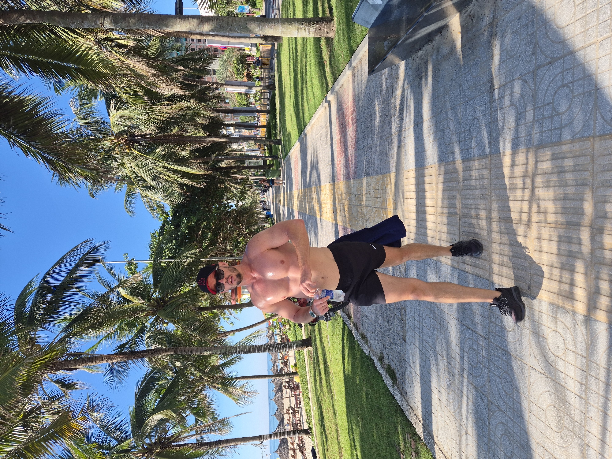 A shirtless man in black shorts, a baseball cap, and sunglasses is jogging along a paved walkway lined with palm trees. He is holding a bottle of water in his right hand. To his left is a grassy area with beach chairs and umbrellas. In the background, there are buildings and the ocean. The scene appears to be the beach promenade in Đà Nẵng, Vietnam.