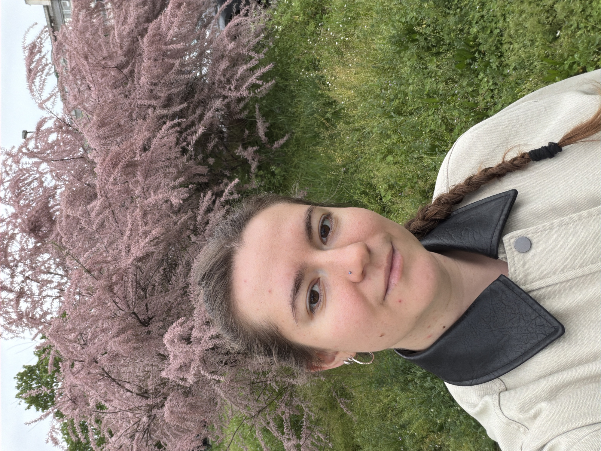 A portrait-oriented photo captures a young woman from the chest up, facing the viewer with a slight, pleasant smile. She has dark brown eyes, light brown hair styled in a braid over her left shoulder secured with a black hair tie, and a small stud piercing on her right nostril. She is wearing a light-colored, possibly beige, jacket with a prominent black collar.
The background is an outdoor setting, primarily filled with natural elements. A large, dense bush or tree covered in delicate pink-purple blossoms or leaves dominates the left side of the frame, creating a soft, textured backdrop. Below and to the right of the flowering plant, an expanse of green grass is visible, dotted with tiny white and purple flowers. A building and a light, possibly overcast, sky can be seen partially in the upper background, behind the pink foliage. The scene takes place during the daytime under natural light. There is no visible text.