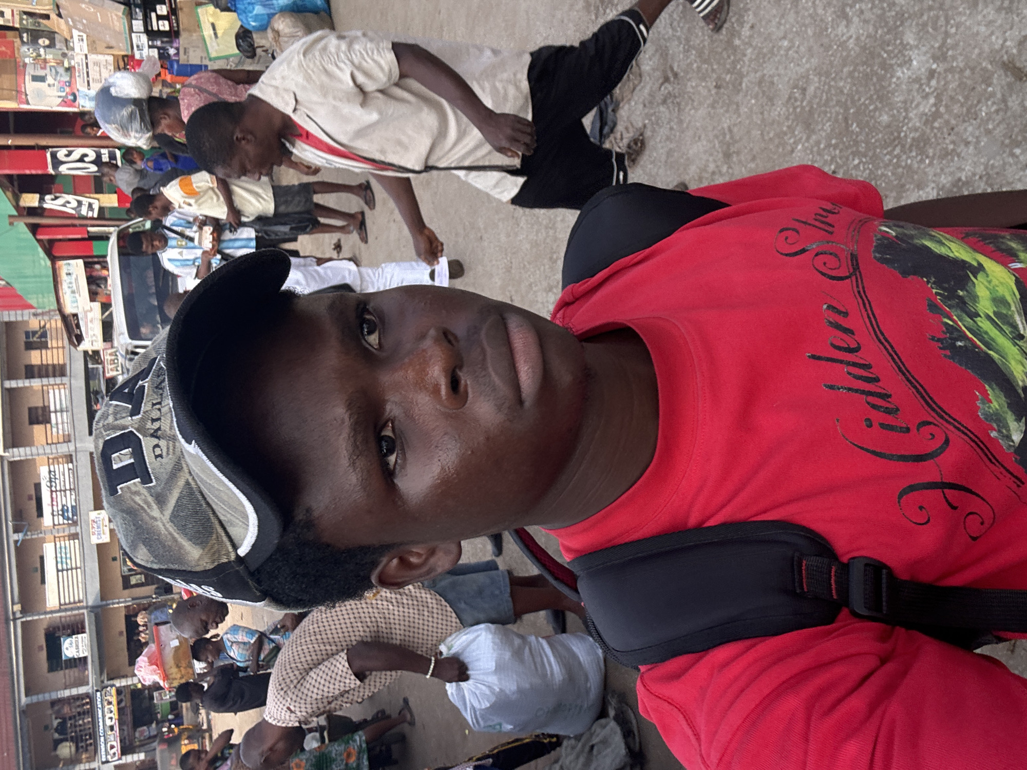 The image depicts a bustling street scene in Onitsha, Nigeria. In the foreground, a young Black male wearing a red t-shirt and a backpack is the primary subject. He is looking towards the camera, with part of his face visible. Behind him, the street is filled with people and stalls. A white van is parked in the background, and several buildings line the street, suggesting a market or commercial area. People are seen carrying various items, including bags of goods. The overall atmosphere appears lively and busy, characteristic of a typical Nigerian street market.