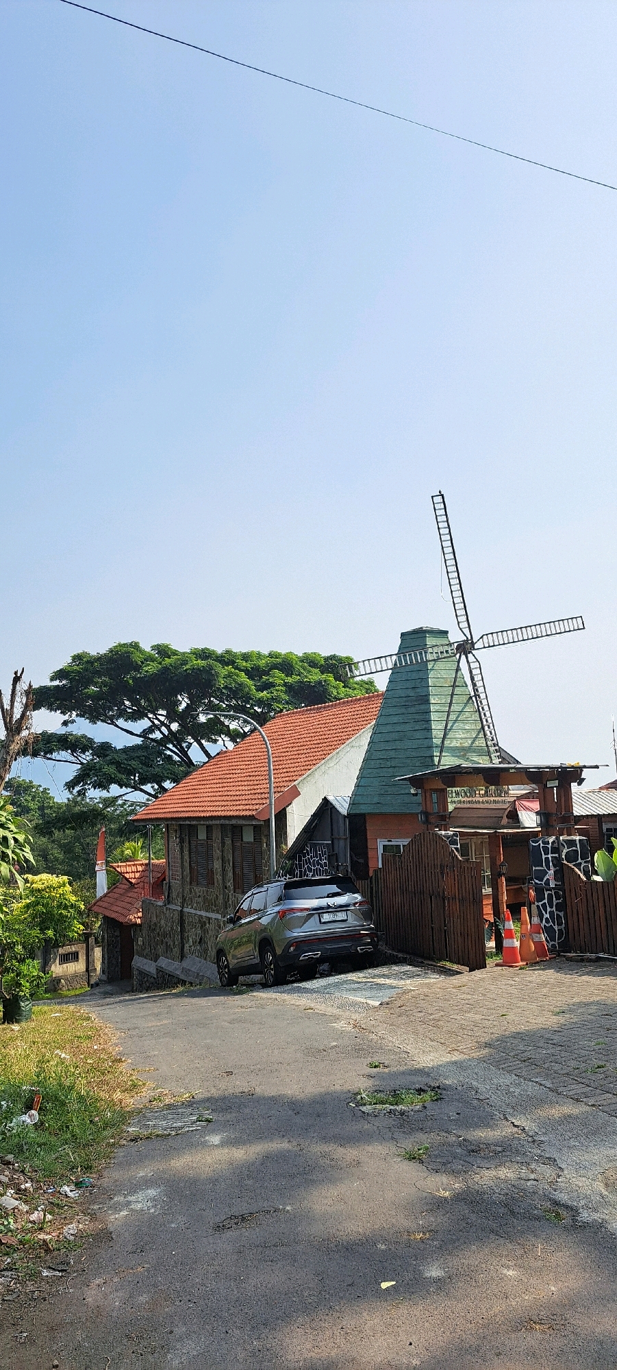 The image depicts a street scene in Prigen, Indonesia, featuring a Dutch-style windmill structure. A gray SUV is parked on the side of a paved road that leads towards the windmill building. The building itself has a red tiled roof and a green, conical upper section where the windmill is situated. A wooden fence encloses part of the property, with orange traffic cones visible near the entrance. Lush green trees and foliage surround the structures, and the sky is clear blue. The road surface shows signs of wear and tear, with some patches of grass growing through cracks. The overall impression is that of a roadside attraction or a commercial establishment in a rural or semi-rural setting.