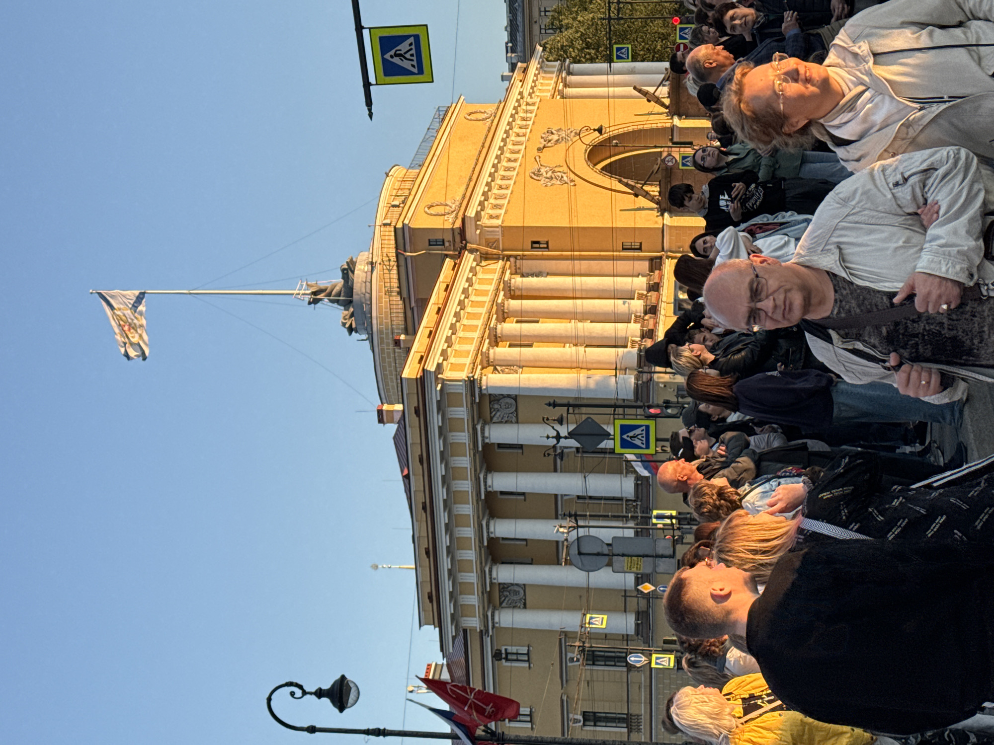 This image captures a crowd of people gathered in front of the Admiralty building in Saint Petersburg, Russia. The sun is low in the sky, casting a warm golden light on the neoclassical architecture of the building and illuminating the faces of the people.
The Admiralty building, a prominent landmark, features a central tower topped with a spire and a flag. The building's facade is characterized by a colonnade of white columns and ornate detailing. Multiple traffic and pedestrian crossing signs are visible, indicating a public square or intersection.
The crowd is diverse, with individuals of various ages and ethnicities. People are standing, walking, and interacting, with some looking towards the building or the spire, suggesting they are observing an event or taking in the scenery. Clothing styles range from casual to slightly more formal, reflecting the time of day and the occasion.
Several flags are displayed on poles in the foreground, including the Russian flag and what appears to be a naval ensign or a historical Russian flag. A street lamp with an intricate design stands to the left of the frame.
The overall atmosphere seems to be one of a public gathering, possibly during a festival, commemoration, or simply a popular tourist spot. The clear blue sky suggests fair weather.