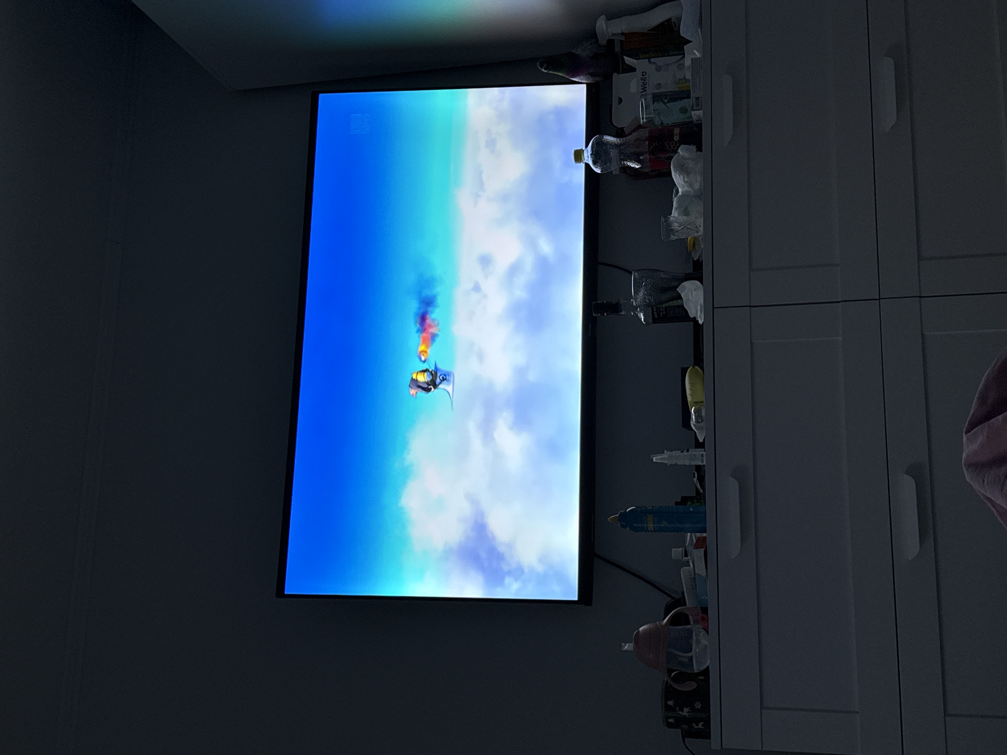The image captures a dimly lit room in Moscow, Russia, featuring a large flat-screen television. The TV screen, which appears vertically oriented within the image frame, displays a vibrant animated scene. This scene depicts a character, seemingly in a flying suit or jetpack, soaring through a bright blue sky dotted with white clouds. The character leaves a colorful trail of red, orange, blue, and green smoke. Faint white text or symbols, possibly channel branding, are visible in the top left corner of the screen.
To the right of the television, a light-colored cabinet or set of drawers is visible. Its top surface is cluttered with various household items, including several plastic bottles (one appearing to be a blue sports bottle, another a pink baby bottle), jars, and white product packaging. The text "Wella" and "fine hair" can be clearly read on one of these packages.
The overall dimness of the room suggests low ambient light, making the bright TV screen the dominant light source. A subtle, rainbow-like light reflection is present on the upper left edge of the screen and adjacent wall. No individuals are directly visible in the room.
