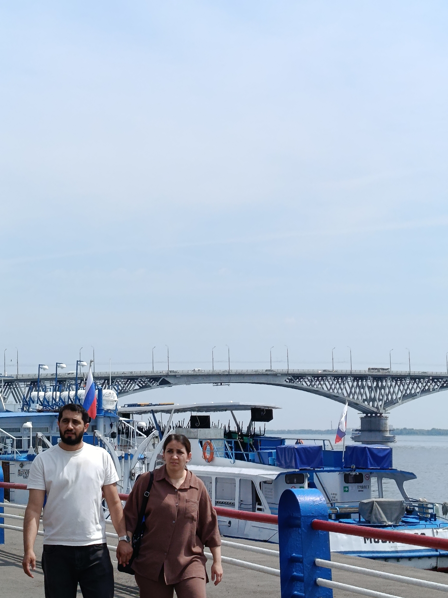 The image depicts a couple walking hand-in-hand along a waterfront promenade in Saratov, Russia. The couple consists of a man with a beard wearing a white t-shirt and black pants, and a woman in a brown button-down shirt and pants. Behind them, several boats are docked, one prominently displaying a Russian flag. In the background, a large, multi-span bridge, likely the Saratov Bridge, crosses a body of water. The sky is a pale blue with some scattered clouds. The overall scene suggests a leisurely stroll on a sunny day by the Volga River.