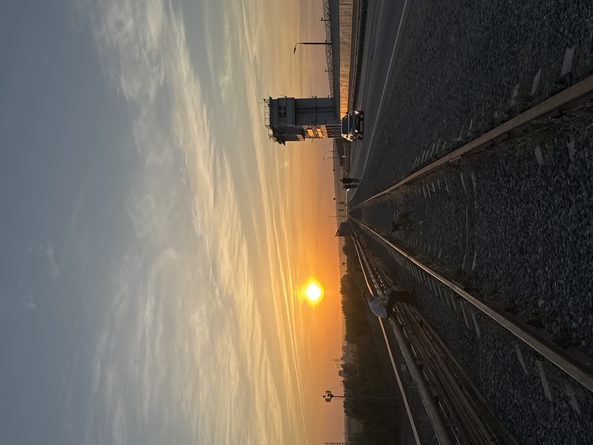 The image captures a sunset scene in Chaikovsky, Russia, with the sun positioned low in the sky, casting an orange glow. A man is perched on a railway track. Parallel to the tracks, a road runs alongside, featuring a building and a car. In the background, a bridge structure is visible against the sky, partially obscured by the sunset's haze. Contrails stretch across the sky. The overall composition suggests a moment captured during the evening.