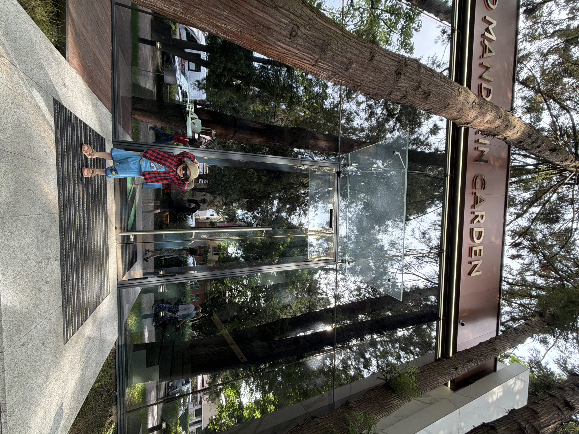 The image shows a child in a red plaid shirt, denim shorts, sandals, and sunglasses standing at the entrance of a building in Sochi, Russia. The building is a modern structure with a large glass facade, which reflects the surrounding trees and sky. A tall sign next to the entrance reads "MANDARIN GARDEN" in gold lettering. A tree trunk partially obscures the view in the upper part of the image. The child appears to be posing for a photo, holding their hand up to their head as if shielding their eyes. The background reflects the urban environment of Sochi, with hints of other buildings and vehicles visible in the glass.