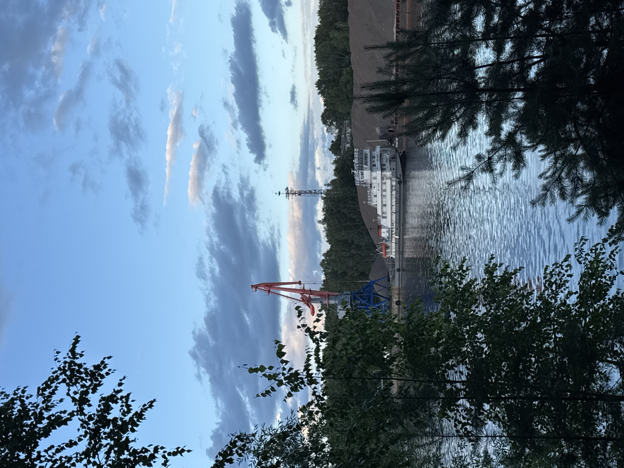 The image depicts a waterfront scene in Chaikovsky, Russia, likely in the late afternoon or early evening given the soft, diffused light and the dramatic cloud formations. The sky is a vibrant blue, streaked with wispy white and darker gray clouds, creating a dynamic visual effect.
Dominating the water is a white vessel, appearing to be a ship or a large boat, docked at a pier. The ship has multiple decks and windows, suggesting it is a passenger vessel or a working ship. In the background, a large industrial crane with red and blue components stands tall, hinting at port activities or construction. Behind the crane and the ship, a dense line of green trees forms a natural backdrop, contrasting with the industrial elements. Further in the distance, a large mound of dark material, possibly coal or industrial waste, is visible.
In the foreground, dark green foliage of trees frames the scene, with some branches extending into the frame, adding depth and a natural element to the composition. The water itself is calm but rippled, reflecting the light of the sky and the surrounding structures. The overall atmosphere is one of a working industrial waterfront bathed in the serene light of dusk. No people are visible in the image. No discernible text is present.