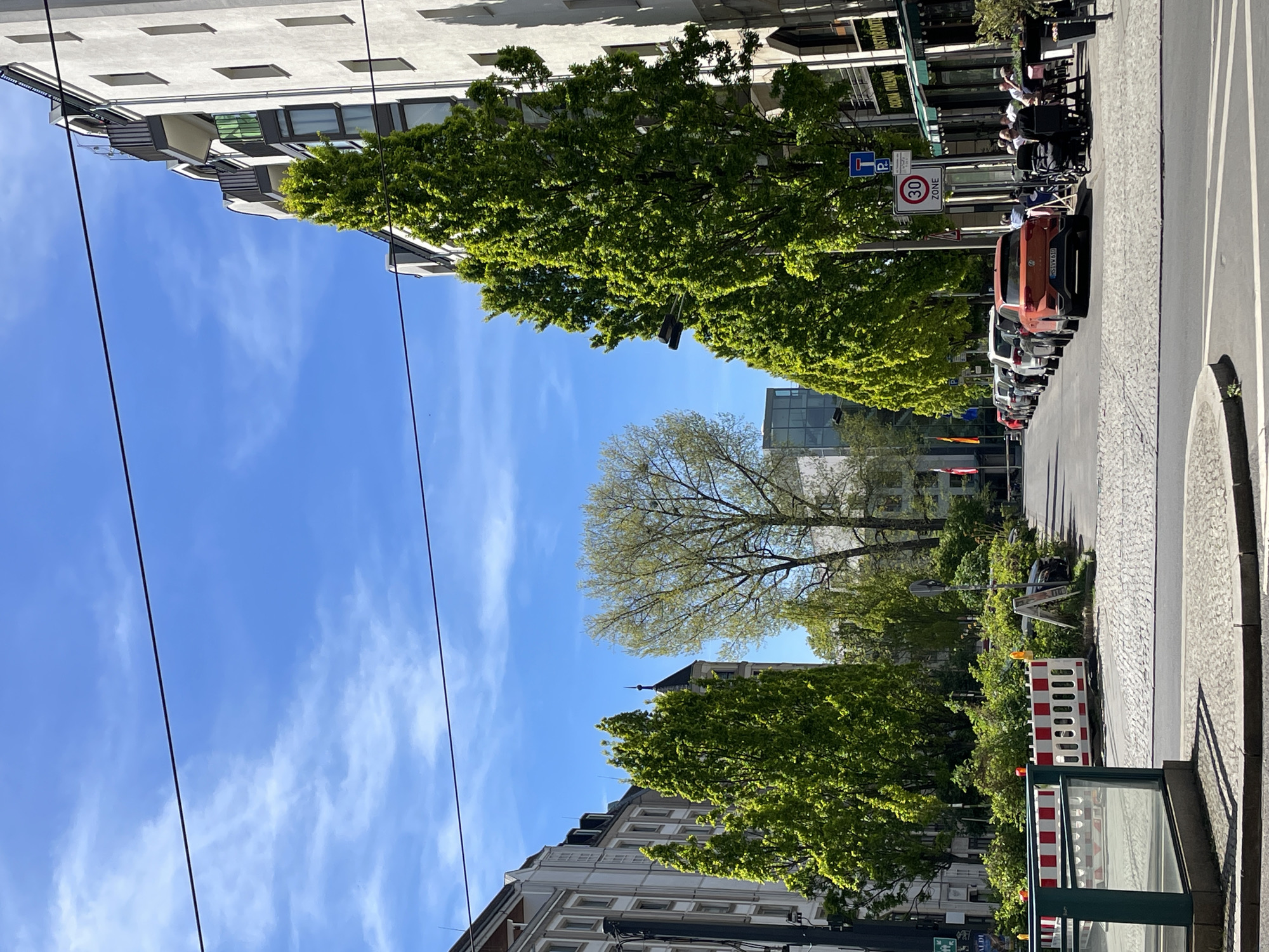 The image captures a lively daytime street scene in Berlin, Germany, under a clear blue sky with sparse white clouds. The view looks down a city street flanked by multi-story buildings and abundant greenery.
On the left side, modern buildings with numerous windows and balconies are partially hidden by large, verdant trees, suggesting spring or early summer. Overhead wires crisscross the sky.
The right side of the image features a cobblestone sidewalk and an asphalt road. A line of cars is parked, including a prominent orange SUV/crossover with a Berlin license plate, "B SY 630". Adjacent to the parked cars, several people are visible seated at outdoor tables, dining or socializing at a restaurant. A green awning bears the partial text "RISTORANTE ITALIANA".
Street signage is evident, including a circular "ZONE 30" speed limit sign and a blue parking sign with a white "P". Towards the bottom right, red and white striped construction barriers are set up, along with a dark green kiosk-like structure. In the distance, a building with a distinctive dark, pointed roof or spire is visible above the tree line. The bright sunshine indicates pleasant weather, conducive to outdoor dining and city life.