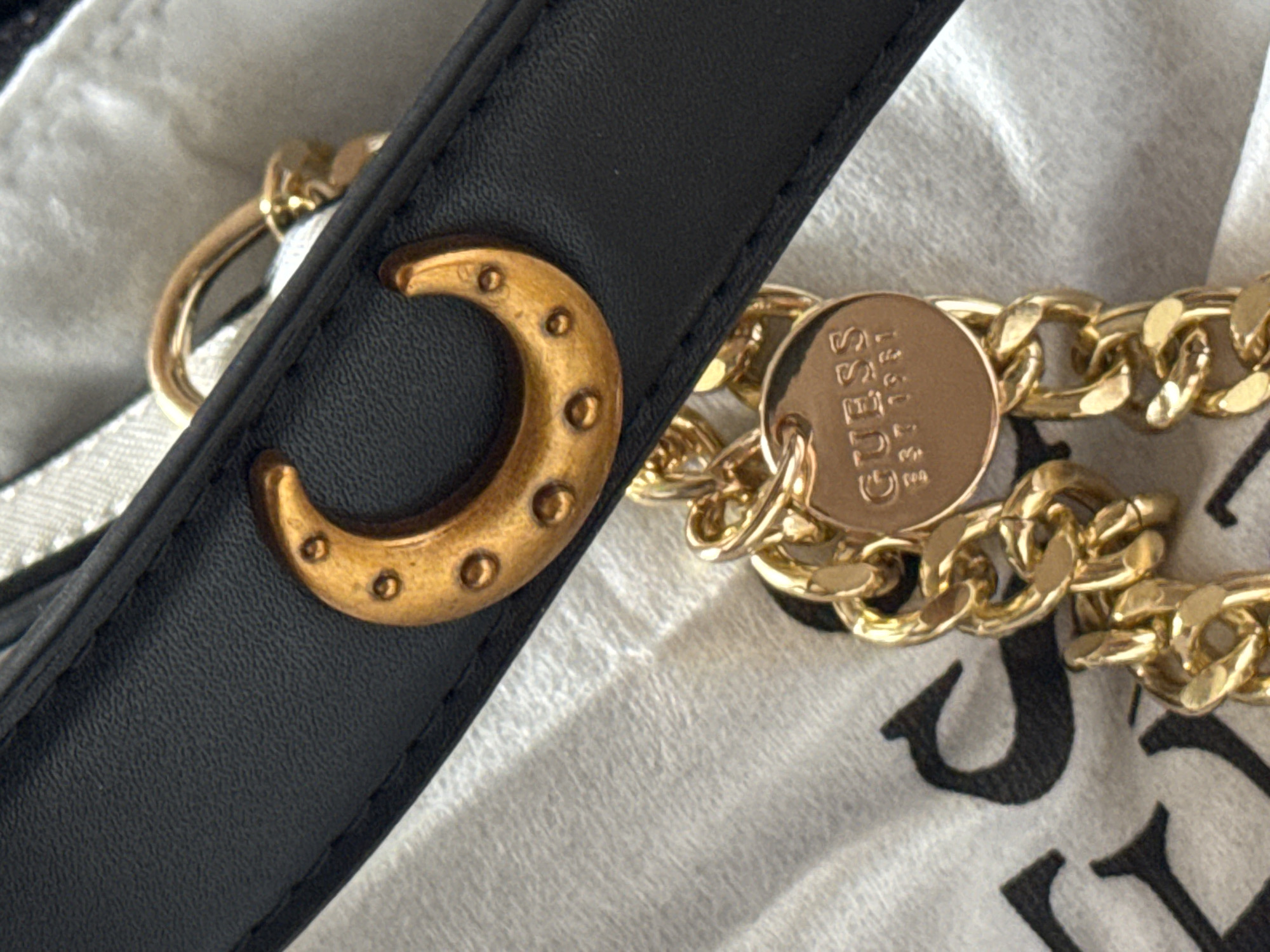 The image shows a close-up of a black leather strap with gold-toned hardware. A circular gold charm with the embossed text "GUESS EST 1981" is attached to a chunky gold chain. Another gold embellishment, shaped like a crescent moon with small circular accents, is also attached to the leather strap. The background consists of a textured white fabric, possibly part of a bag or clothing. The lighting suggests an indoor setting, possibly with artificial light, as there are highlights on the metal components. There are no people visible, and no specific time of day or weather conditions can be determined. The location context of Izhevsk, Russia, is provided, but there are no visual cues in the image to confirm this. The overall impression is one of a fashion accessory detail.