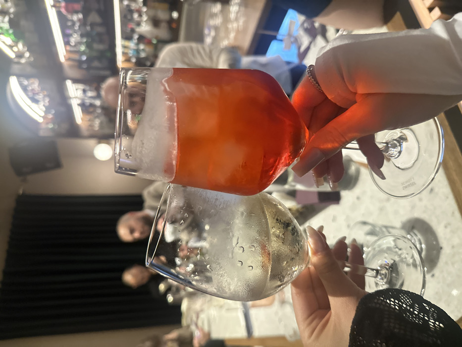 The image displays an indoor scene within a bar or restaurant setting. In the foreground, two hands are observed holding stemmed glasses in a probable toast. The left hand, partially obscured by a dark, possibly lace or textured garment sleeve, grasps a clear glass containing a pale yellow or clear liquid with ice cubes and external condensation. The right hand, wearing a light-colored long-sleeved garment, holds a clear glass filled with a vibrant orange-red cocktail, also with ice cubes and condensation; a thin ring adorned with small stones is visible on its ring finger. Both hands feature long, manicured, light-colored fingernails.
In the blurred midground, several individuals are present. A prominent male figure with a bald or closely shaven head and a beard, wearing a light-colored top, is visible towards the left. Other indistinct figures are present further back. The background features dark, heavy curtains on the left and a brightly lit, well-stocked bar on the right, displaying numerous bottles arranged on shelves, illuminated from above and behind. The overall ambiance is dimly lit, characteristic of an evening establishment. The specific location within Yerevan, Armenia, is not discernible from the visual evidence.