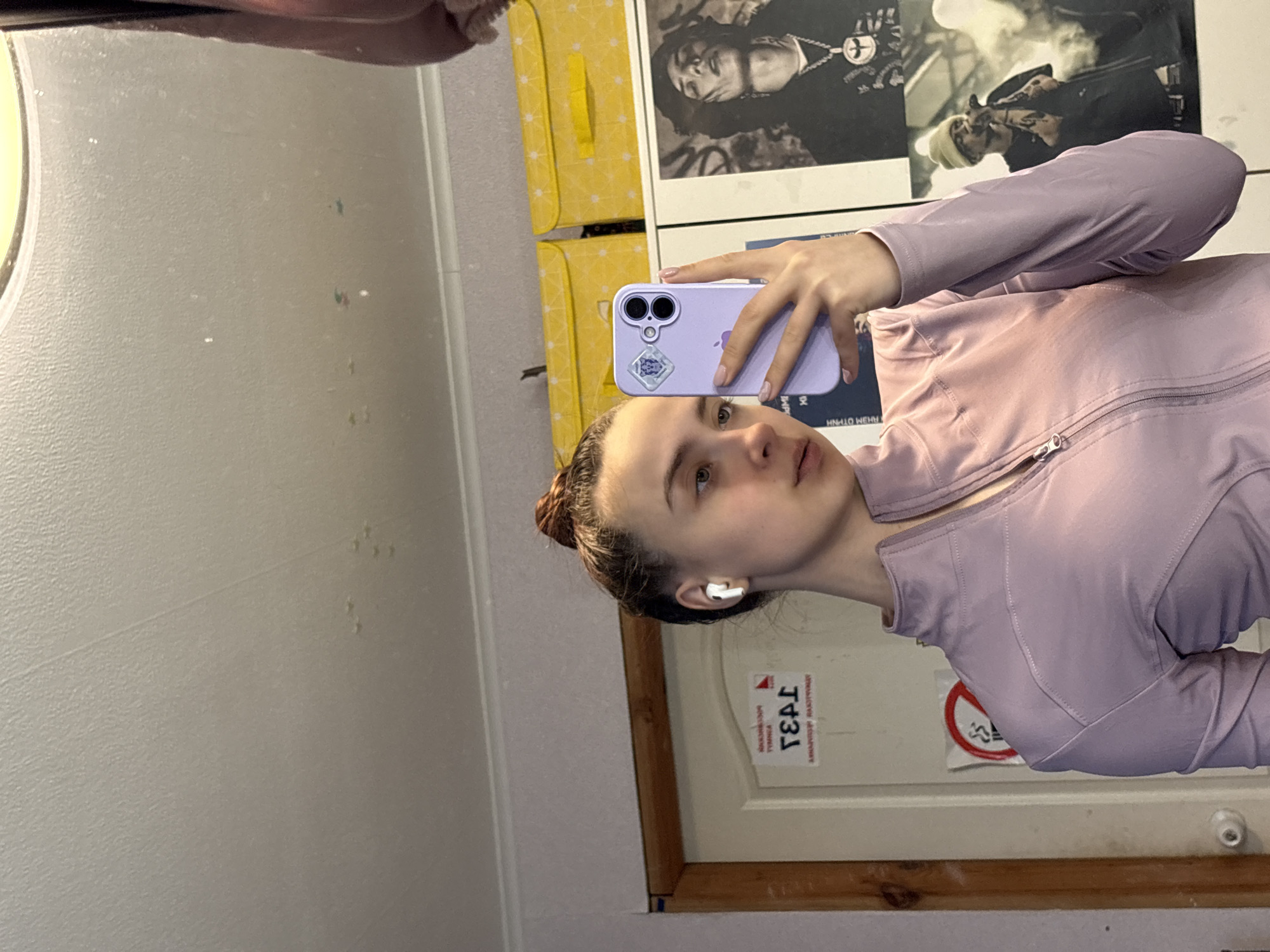 A young woman is captured in a mirror selfie, taken indoors in what appears to be a room in Prikamskiy, Russia. She is positioned slightly off-center, holding a lavender-colored iPhone with a distinctive dual-camera setup and a small sticker on its back, obscuring part of her face. She is wearing a light purple, long-sleeved athletic jacket with a visible zipper and textured seams. A white wireless earbud is present in her left ear, and her dark hair is pulled back, likely in a bun. Her expression is neutral, focused on the act of taking the picture.
The background, visible through the mirror's reflection, includes a plain, light-colored wall. To the left, a bright yellow object with a dotted pattern, possibly a storage box or piece of furniture, is visible. On the wall behind her, several posters are displayed; one clearly shows a person with dreadlocks and a necklace, while another depicts a person wearing a beanie, suggesting music or pop culture themes. Below the posters, a wooden frame, possibly part of a door or the mirror itself, is visible. Affixed to this frame are reversed stickers: one reads "ТЕАТР" (Russian for "Theater") and likely "ВЛАДИМИР" below it, and another is a "no smoking" symbol (a red circle with a crossed-out cigarette). The mirror surface itself shows some minor smudges or dust particles.