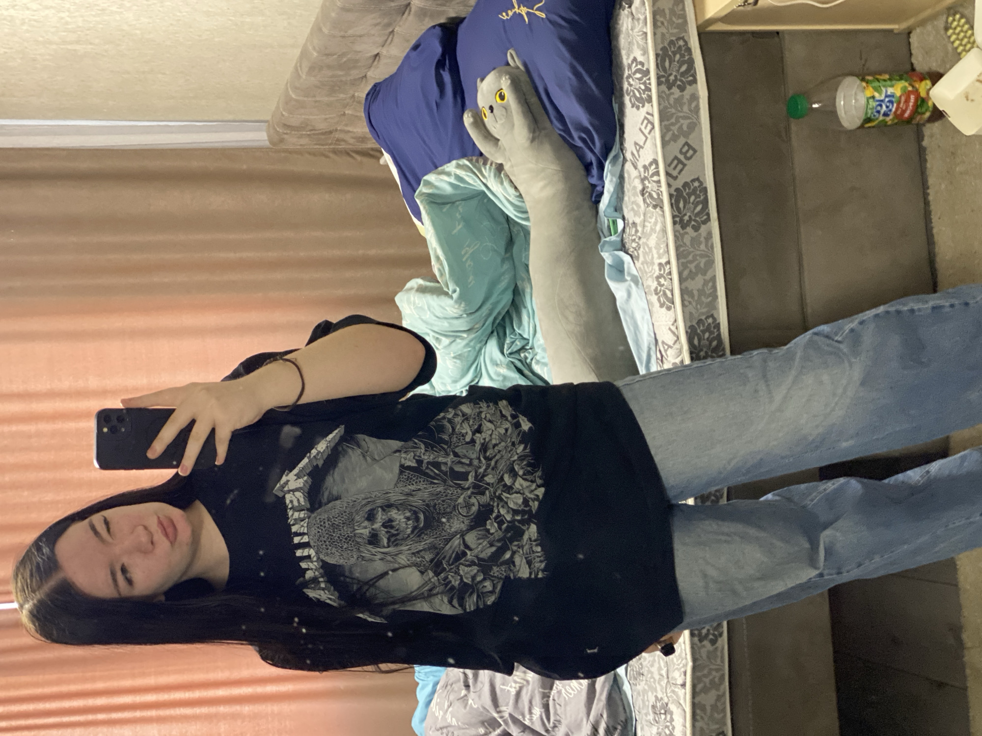A woman is taking a selfie in a bedroom. She is wearing a black t-shirt with a large graphic design and light blue jeans. She is holding a black smartphone in her left hand. The bedroom contains a bed with grey bedding and a blue pillow, as well as a long grey stuffed cat toy. A juice bottle is visible on the floor next to the bed. The scene is likely taking place indoors, in a private residence. There is no indication of the location being in the city of Ternopil, Ukraine.