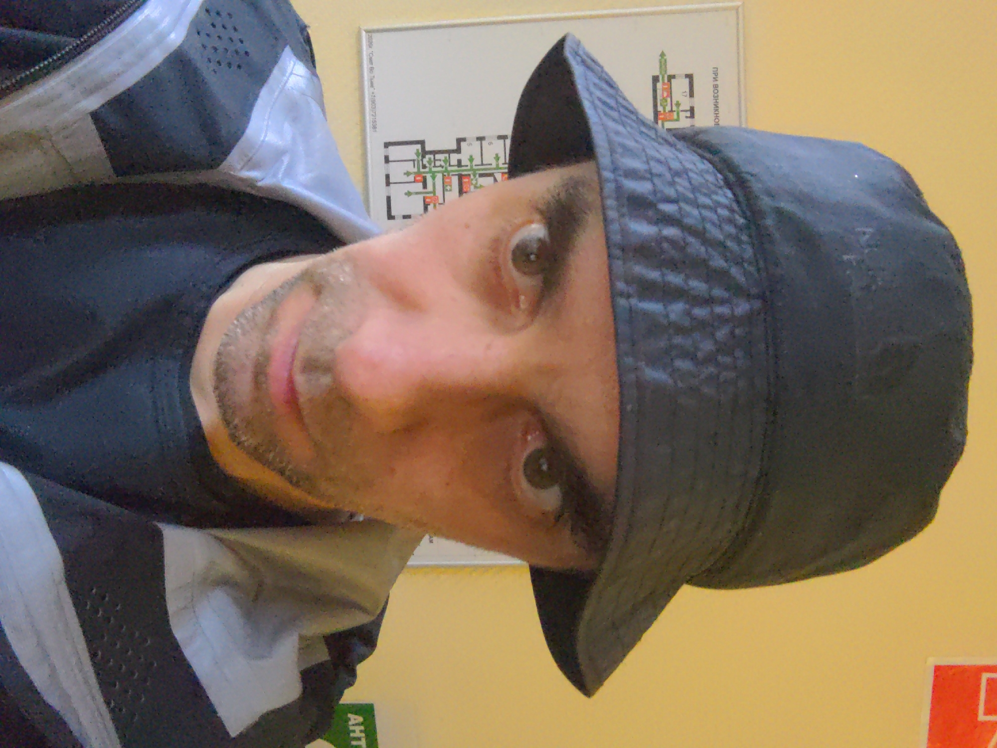 A selfie-style portrait shows a light-skinned male with brown eyes, a trimmed beard, and a black bucket hat. He wears a black shirt and a gray and black jacket. Behind him is a schematic diagram mounted on a pale yellow wall. The text on the diagram includes the Russian phrase "ПРИ ВОЗНИКНОВЕНИИ". A green sign with text in Cyrillic is visible in the lower left corner, and a red sign is partially visible in the lower right. The location appears to be inside a building, likely in Russia, given the Cyrillic writing. The city is unknown.