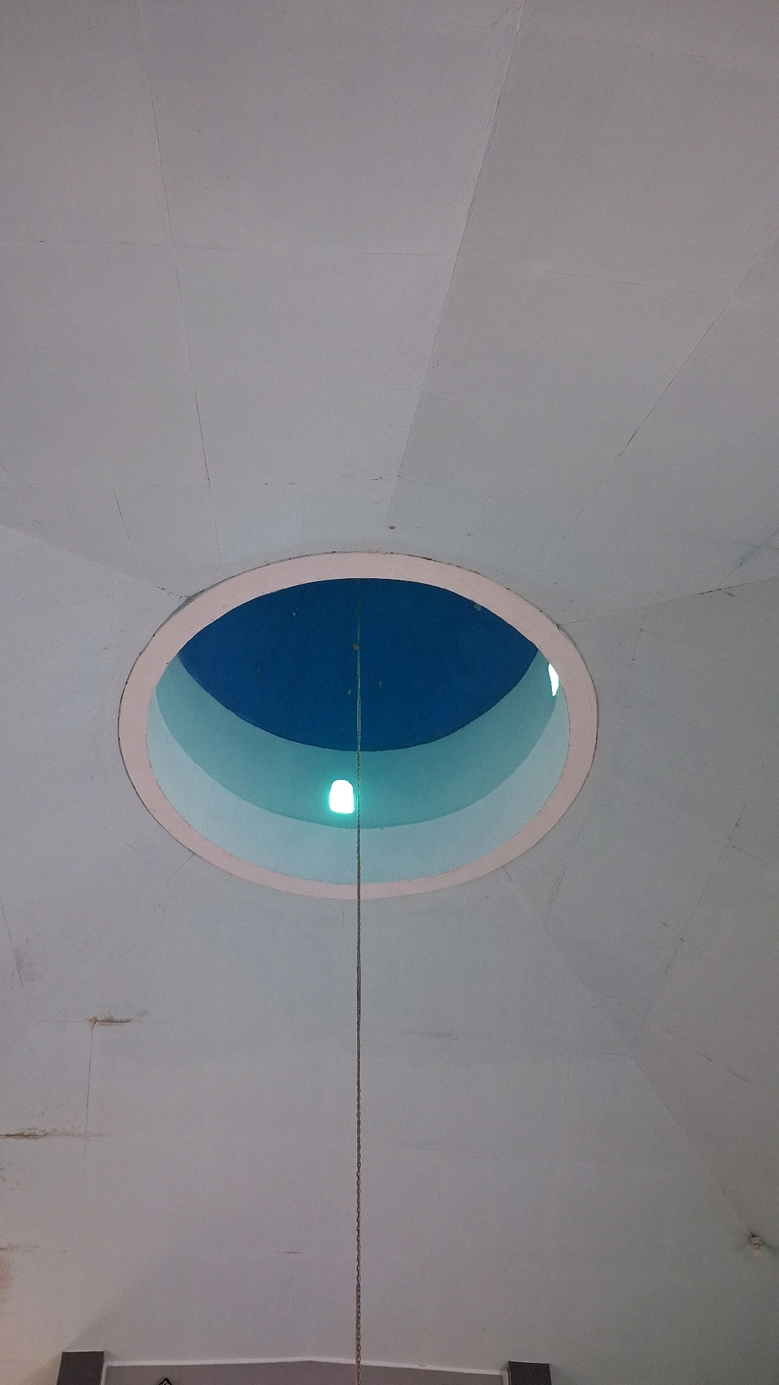 A low-angle shot looks up at a domed ceiling with a central circular opening. The ceiling is painted a pale gray and appears to be constructed from panels. The opening in the ceiling reveals layers of curved surfaces in shades of blue and light blue, leading to a deep blue interior. A thin metal chain hangs vertically through the center of the opening, extending from above into the visible space. A small, bright white light source is visible within the opening, illuminating the chain and the lower layers of the curved interior. The overall impression is of an architectural feature, possibly a skylight or a decorative element within a building. The image is located in the city of Giriwarno, Indonesia.
