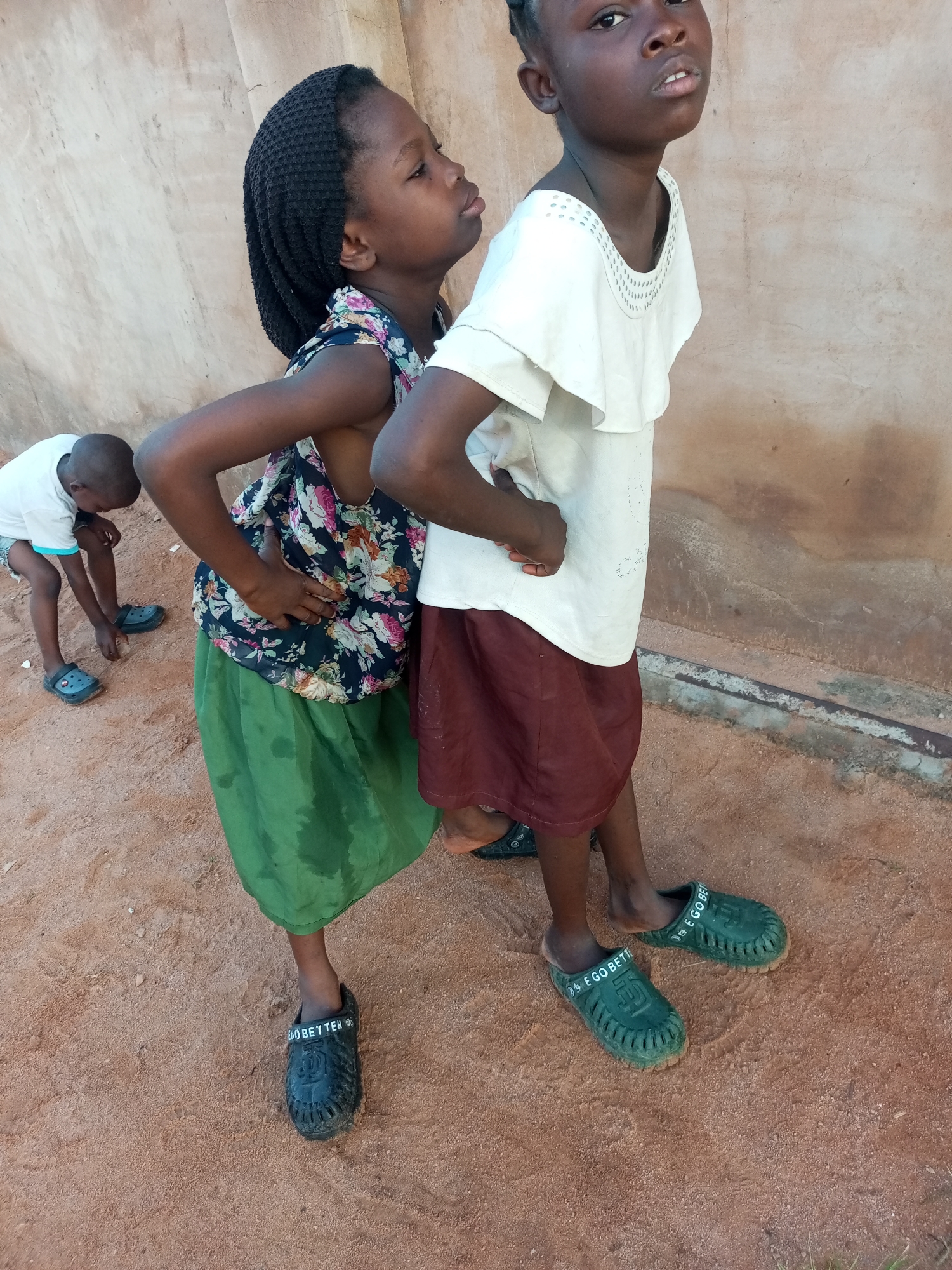 In Tempure, Nigeria, a brown dirt ground is the setting for three children. Two young girls are standing in the foreground, their bodies angled towards the viewer. The girl on the left has braided black hair, wears a floral top and a green skirt, and is facing left with her arm resting on her hip. She is wearing black clogs. The girl on the right is wearing a white ruffled top and a maroon skirt, and she is facing the viewer directly with her hands on her hips. She is wearing green clogs. A younger boy, wearing a white t-shirt and grey shorts, is in the background to the left, bending down on the dirt ground. He is wearing black clogs. The children are positioned in front of a tan, textured wall.