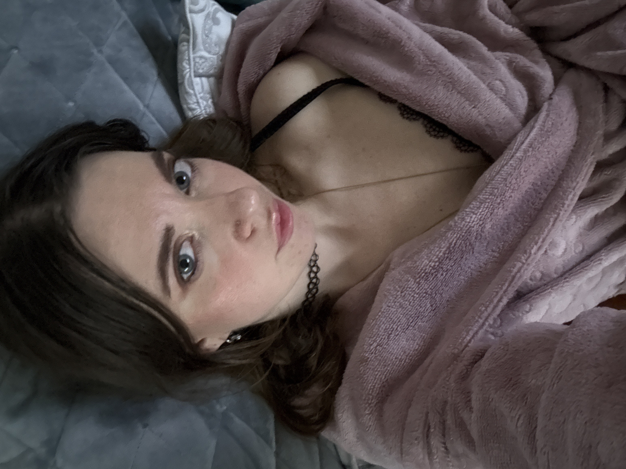 This is a close-up, high-angle, portrait-oriented media file featuring a young woman lying on a grey, quilted surface, possibly a bed. She is wrapped in a soft, lavender-colored robe, with some of her chest and a black lace-trimmed bra visible. A thin gold necklace adorns her neck, and a black choker is also visible. Her face is turned towards the camera, with her eyes wide open and looking directly into the lens. She has light skin, rosy cheeks, and light brown hair that falls around her face. Her lips are slightly parted, revealing a hint of her tongue. The lighting appears to be indoor, with the time of day and weather not discernable. The overall mood seems intimate and personal. There is no visible text in the image. The location context is Chaikovsky, Russia.