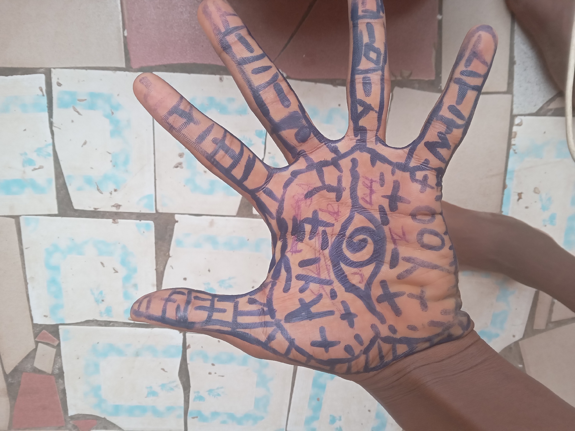 An open human hand, likely that of a child or young person, is captured in a close-up, eye-level shot against a tiled floor, indicative of a setting in Onitsha, Nigeria. The hand's palm and fingers are extensively decorated with dark blue or black marker drawings, forming a dense pattern of abstract lines, crosses, squares, and geometric shapes. A prominent spiral or eye-like symbol is drawn in the center of the palm. On the fingers, especially the index finger, letter-like symbols are visible, with characters resembling "ATHW" or "HITH" discernible on the index finger. The hand's skin tone is dark. The background features a tiled floor composed of a mosaic of light-colored squares, some with faded blue geometric patterns, intermixed with plain beige and reddish-brown tiles, some appearing broken or irregularly shaped. The bright lighting suggests it is daytime. The image depicts a creative activity involving body art.