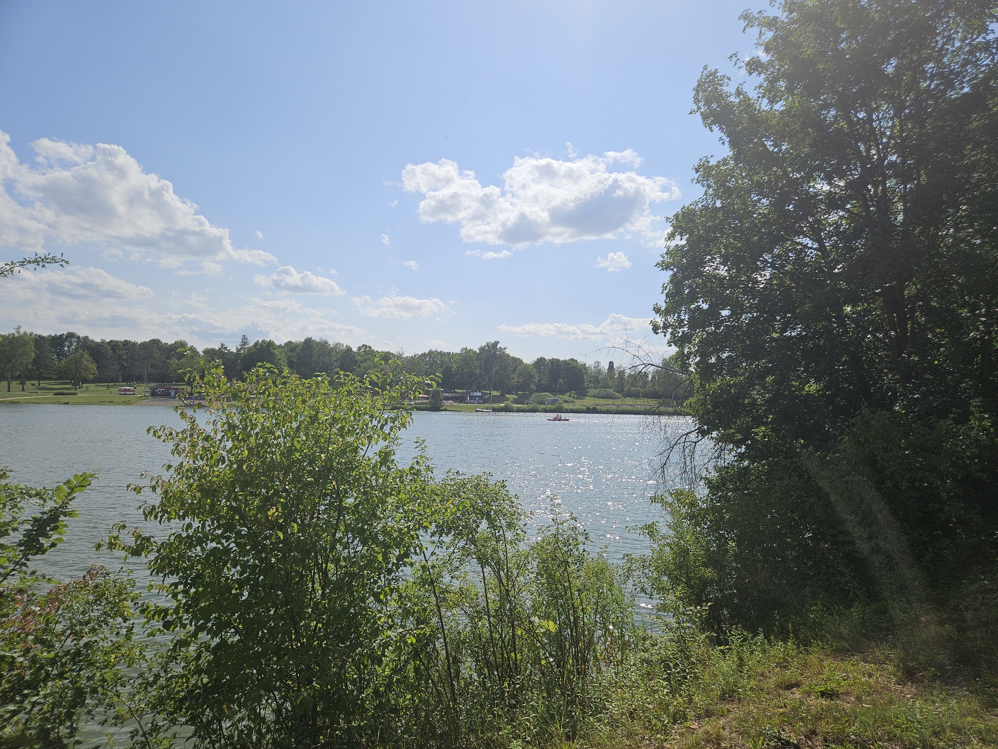 The image depicts a serene lake scene in Rainau, Germany. The foreground is dominated by lush green trees and vegetation, with a body of calm water stretching into the distance. The water reflects the bright, sunlit sky, which is scattered with fluffy white clouds. In the background, a treeline encircles the lake, and a few buildings and vehicles are visible along the shore. A small red boat with two occupants can be seen on the water, suggesting recreational activity. The overall impression is one of a peaceful, natural landscape.