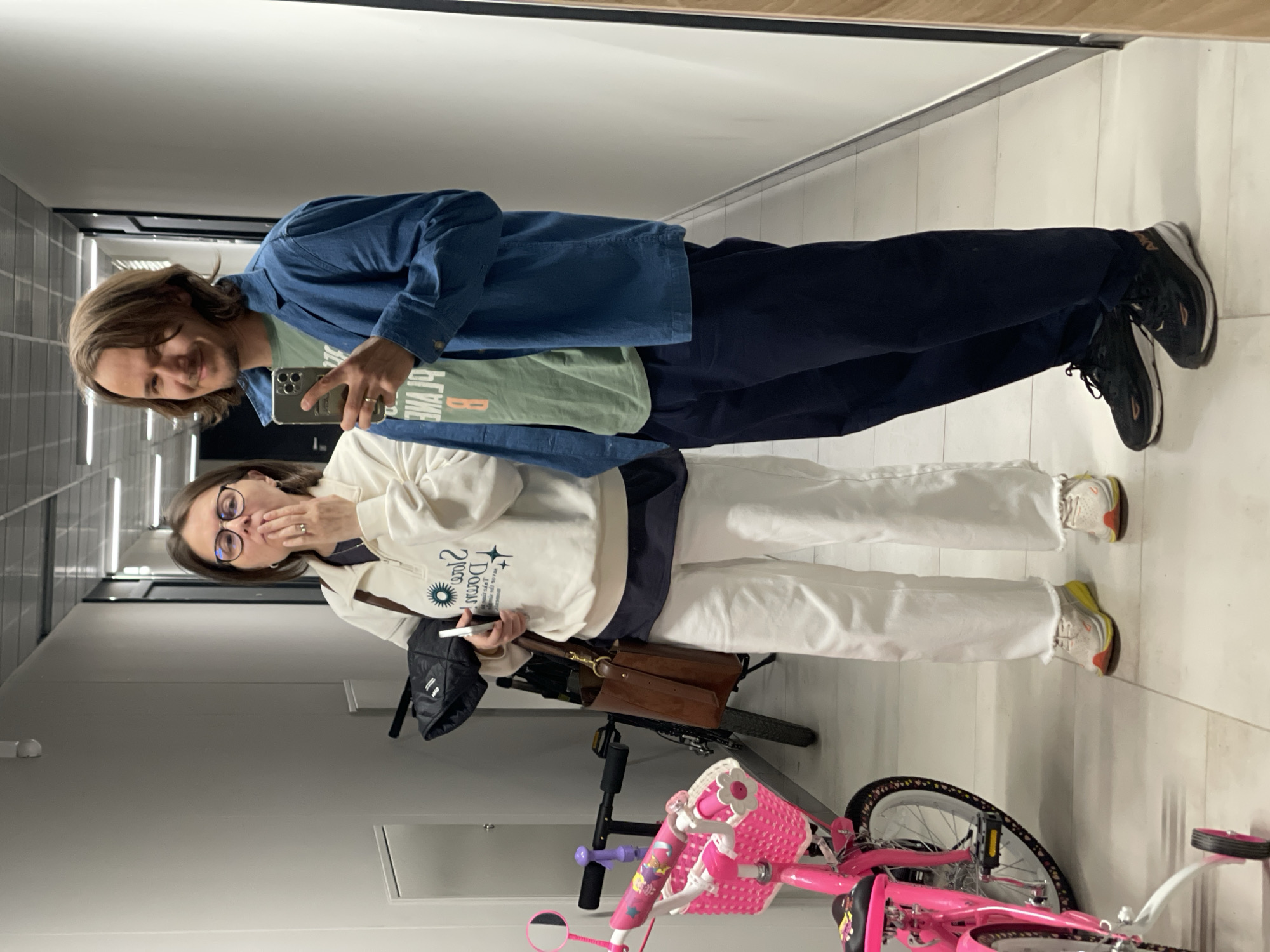 A man and a woman are standing in a hallway with a pink bicycle. The man is on the left, wearing a blue denim jacket, a green t-shirt, dark blue pants, and black sneakers. He is holding a cell phone and taking a picture of himself and the woman in a mirror. The woman is to the right of the man and has glasses. She is wearing a white sweatshirt with the text "Star Dawn" on it and white pants, and white and yellow sneakers. She is holding a cell phone in her right hand and touching her lips with her left. Behind the woman is a brown leather purse, a black shoulder bag, and a black and silver bicycle. The background is a hallway with white walls and a tiled floor.