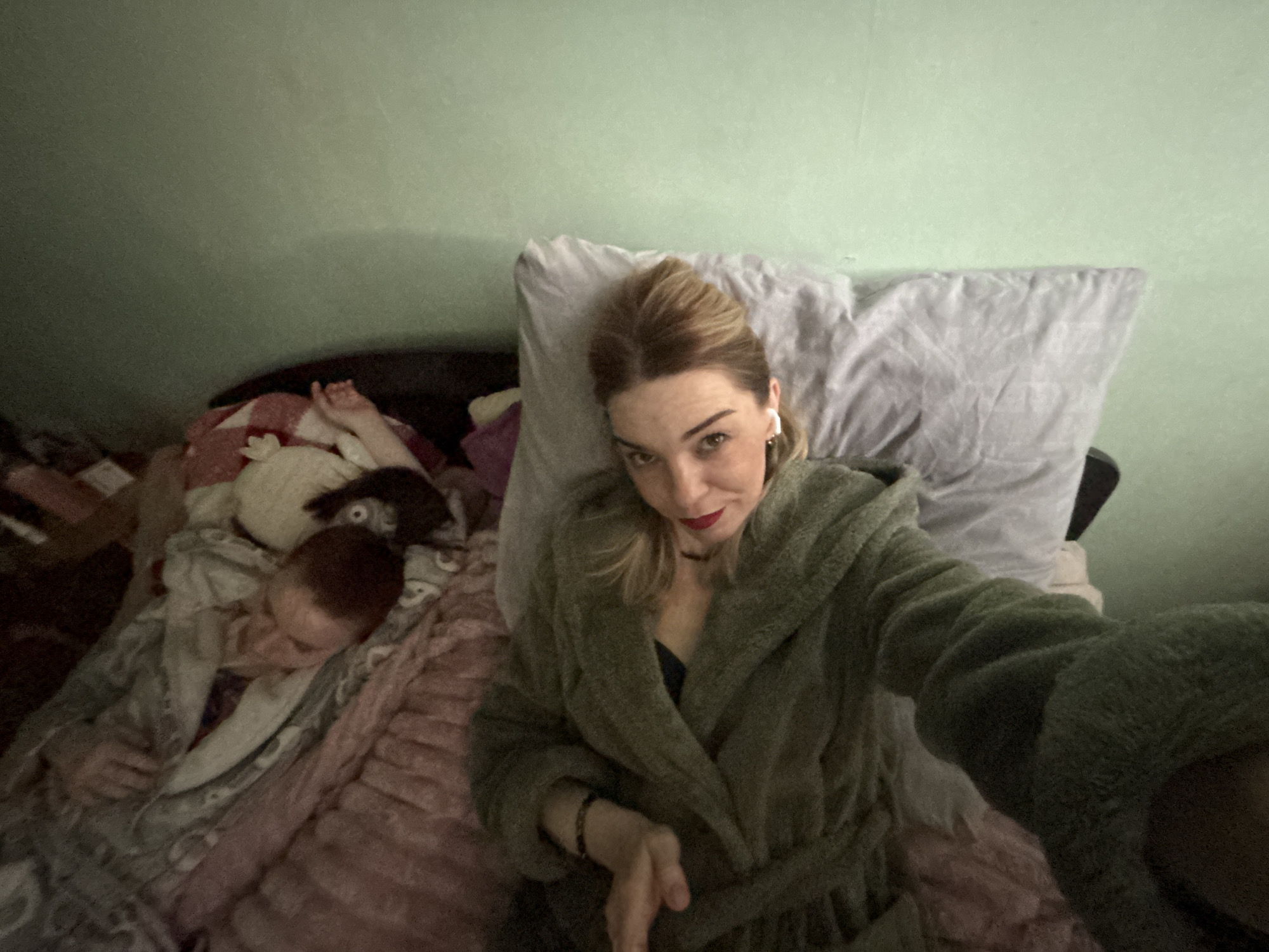 A woman in a green robe is lying on a bed with a gray pillow behind her head. She is wearing wireless earbuds and has red lipstick on. Next to her on the left side of the bed is a child sleeping under a blanket. There are stuffed animals near the child. The wall in the background is light green. The scene appears to be indoors. The location is Murino, Russia.