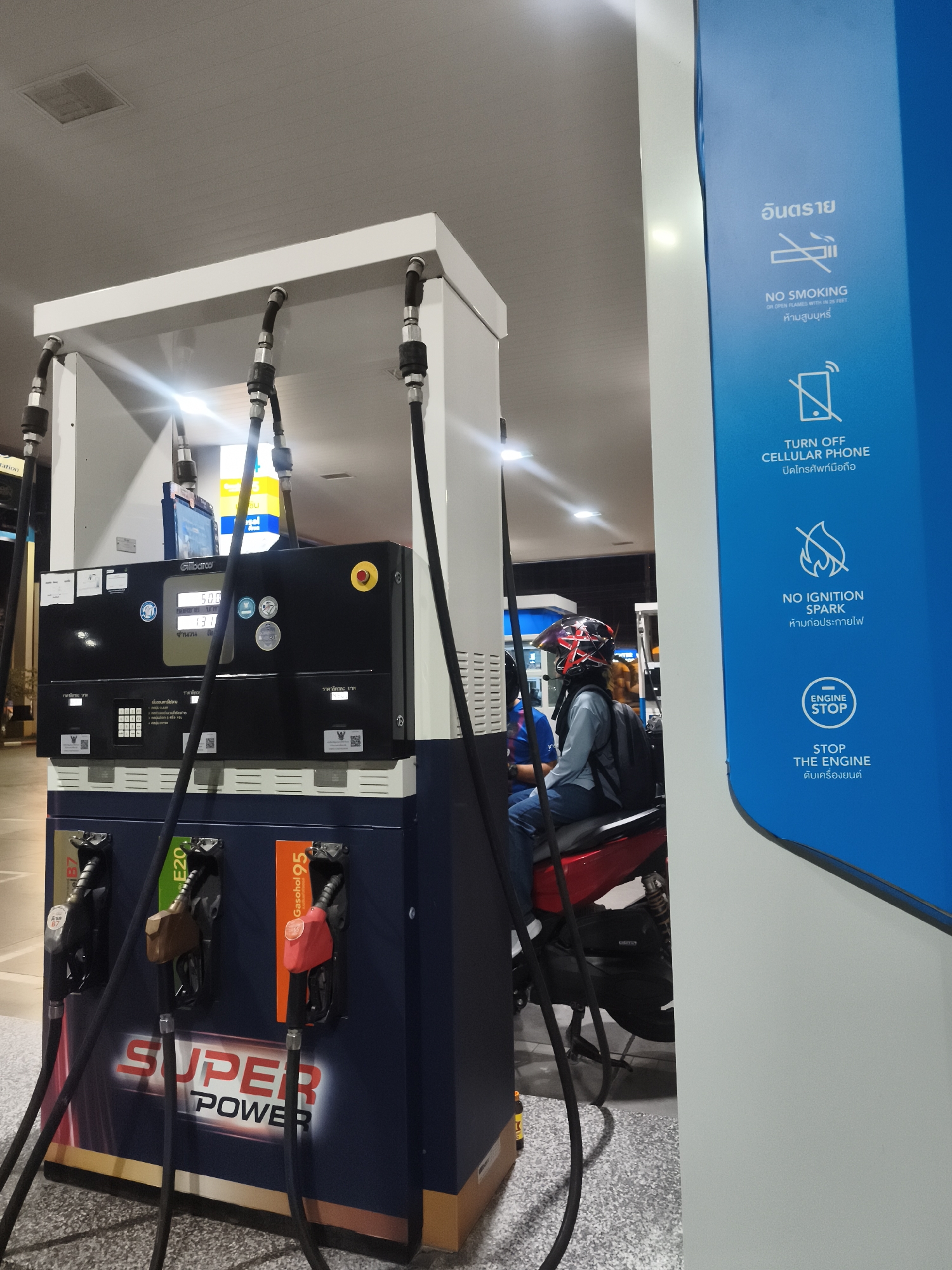 This image depicts a night scene at a gas station in Baan Mae Nam, Thailand. A person wearing a black and red helmet, a light-colored long-sleeve shirt, and a dark backpack is seated on a red motorcycle, positioned next to a multi-product fuel dispenser. The gas pump is dark blue and white, with "SUPER POWER" written prominently on its lower section. It offers three types of fuel, identified by color-coded nozzles and labels: "B7," "E20," and "Gasohol 95" (the red nozzle). The pump's digital display, branded "Gilbarco," shows numbers such as "500" and "131," likely indicating cost and volume.
To the right of the pump, a tall blue sign displays various safety regulations with icons and text in both Thai and English. These include warnings for "อันตราย" (Danger), "NO SMOKING OR OPEN FLAMES WITHIN 25 FEET / ห้ามสูบบุหรี่" (Forbid smoking), "TURN OFF CELLULAR PHONE / ปิดโทรศัพท์มือถือ" (Turn off mobile phone), "NO IGNITION SPARK / ห้ามก่อประกายไฟ" (Forbid sparking), and "ENGINE STOP / STOP THE ENGINE / ดับเครื่องยนต์" (Stop engine). The scene is brightly illuminated by overhead lights, suggesting evening or nighttime activity. The overall setting is functional, with the person likely refueling or waiting to refuel their motorcycle.