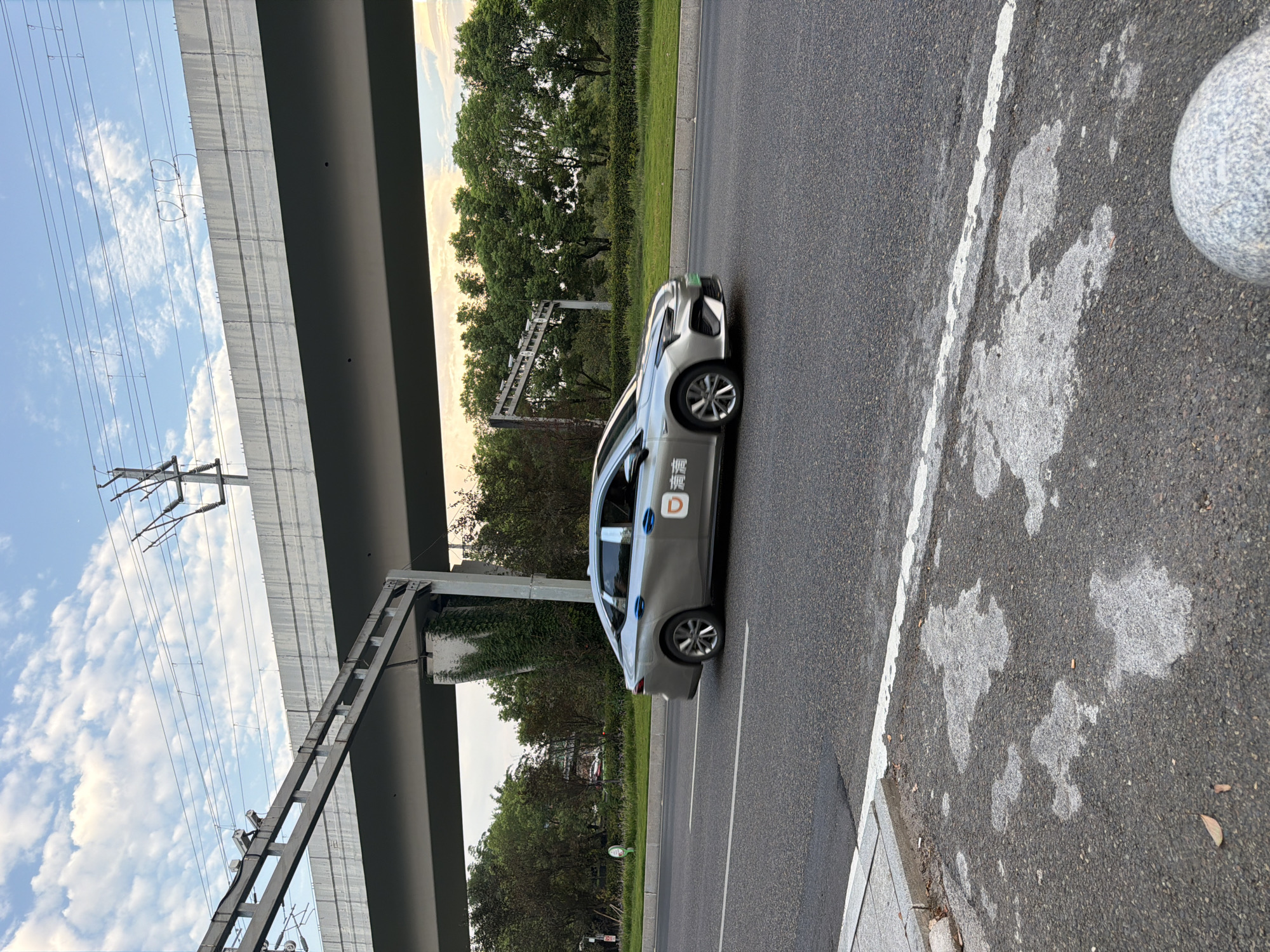 A silver sedan with "DiDi" branding is driving on a road in Jiangjin, China. The car is positioned in the right lane of a multi-lane road. Overhead, a concrete overpass with electrical infrastructure is visible, supported by concrete pillars. Green trees line the side of the road. The sky above is partly cloudy with a soft, diffused light, suggesting it may be late afternoon or early evening. The road surface shows signs of wear, with patches of lighter gray material. A white line demarcates the lanes. In the foreground, a portion of a granite bollard is visible.