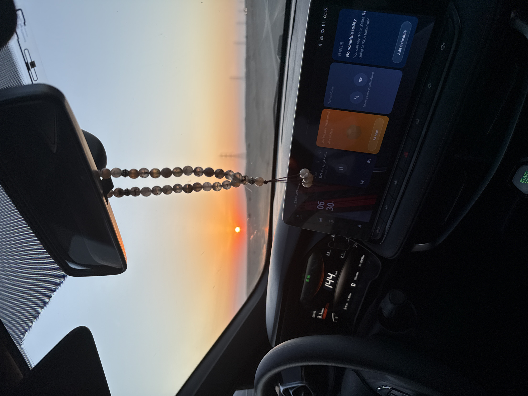 The image is taken from inside a car, looking out through the windshield at a sunset over a flat, desert-like landscape. The sun is a bright orange orb low on the horizon, casting a warm glow across the sky. In the foreground, the car's dashboard is visible, showing a digital speedometer reading 144 km/h. A touchscreen infotainment system is also on display, with various apps and functions visible. A beaded string hangs from the rearview mirror, partially obscuring the view of the sunset. The overall impression is one of a solitary journey, perhaps on a highway or road through a remote area, with the setting sun providing a dramatic backdrop.