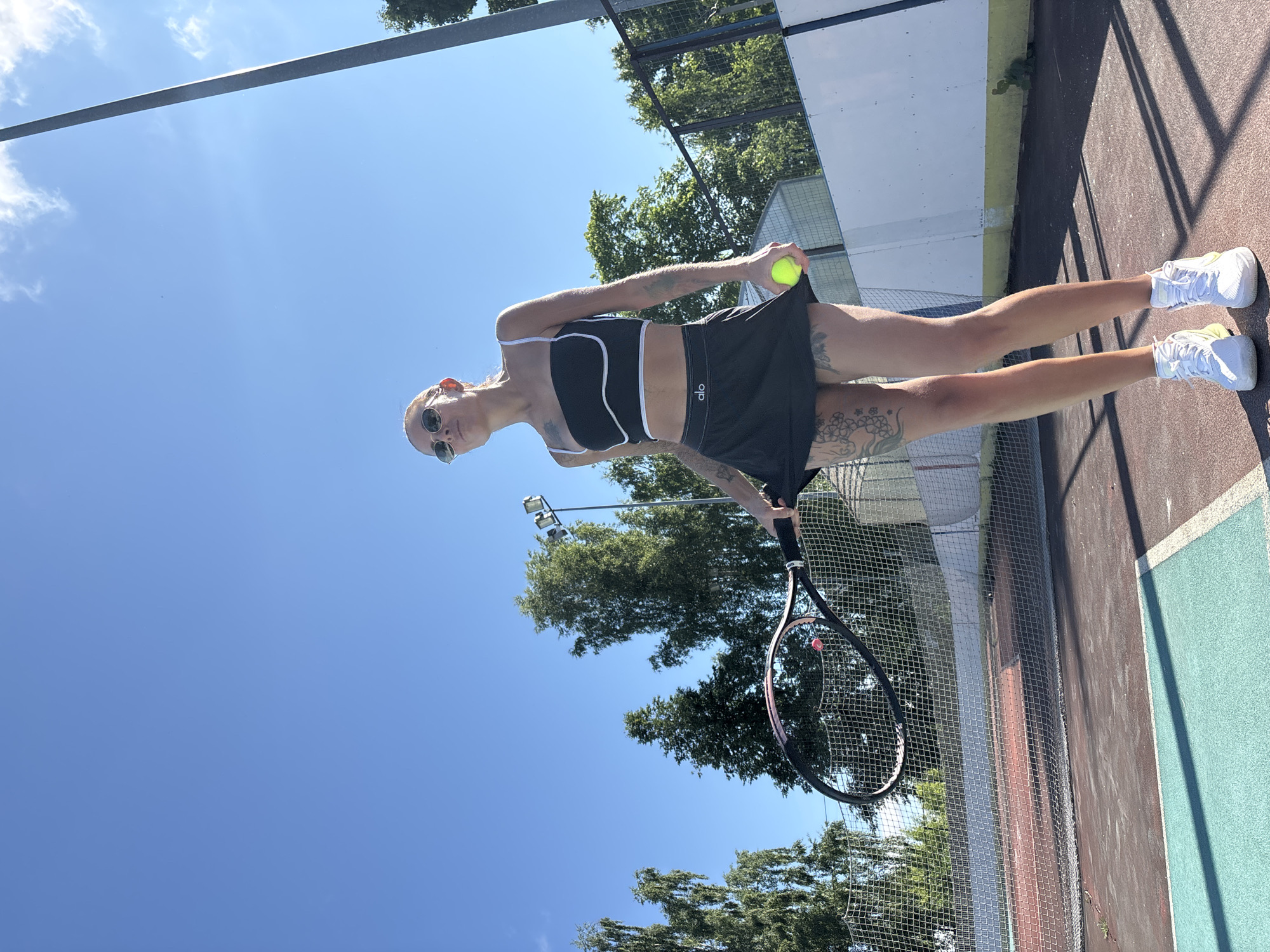 An athletic woman in a black sports bra and skirt holds a tennis racket and a yellow tennis ball on an outdoor tennis court. She wears sunglasses and white sneakers. The court has red and blue surfaces, with a net in the foreground. Trees and a light pole are visible in the background under a clear blue sky. The scene appears to be the city of Chaikovsky, Russia.