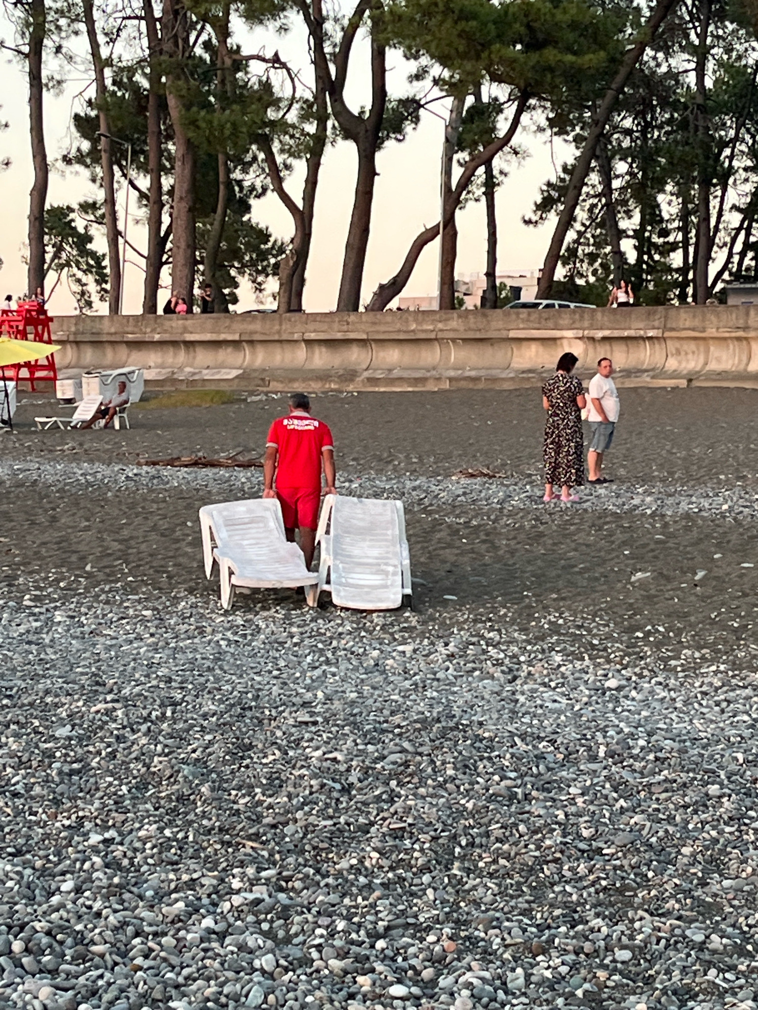 A lifeguard in a red uniform pushes two white lounge chairs on a pebbled beach in Kobuleti, Georgia. In the background, two people, a woman in a patterned dress and a man in a white t-shirt and shorts, stand and observe. Further in the distance, other people are visible sitting on a raised concrete wall topped with trees. The sky is a light, hazy orange, suggesting either dawn or dusk.
