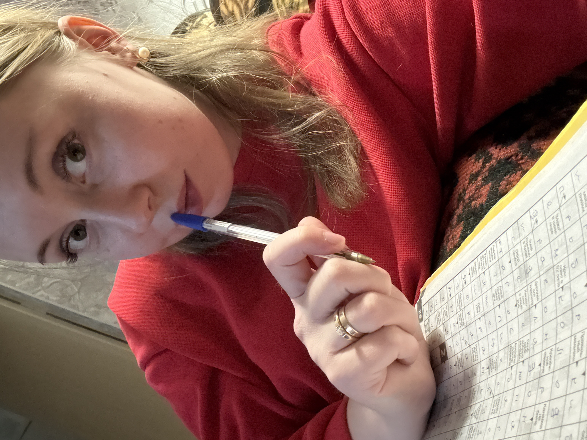 A close-up, slightly tilted image shows a person, likely a woman, from a high angle, focused on her upper body and face. She has light-colored hair and fair skin. Her gaze is directed upwards, suggesting thought or contemplation, while a blue pen is held to her lips. She is wearing a textured red long-sleeved garment, possibly a sweater or sweatshirt. On her left hand, a gold-toned ring, appearing to be two bands with a small stone on one, is visible on her ring finger. A small, pearl-like earring is visible in her right ear.
The person is engaged in an activity that involves a piece of paper with a grid, filled with Cyrillic script and individual letters, typical of a crossword puzzle or word game. The paper has a yellow edge. Next to her, a patterned fabric in dark brown/black and orange/red hues is visible, possibly part of a cushion, blanket, or another piece of furniture. The background is indistinct, appearing as light-colored surfaces indoors. The overall mood suggests quiet concentration.
Visible text on the paper includes numerous Cyrillic letters in grid squares and words like "пиками" (spades), "Межа" (boundary), "пюре" (puree), "Комплект в 52 листа" (Set of 52 sheets), "Дисплей" (Display), "Крепёж изделие" (Fastener product), "обувка" (footwear), and "Канавка болте" (Groove bolt), along with the number "28".
Location context: Unknown, Unknown