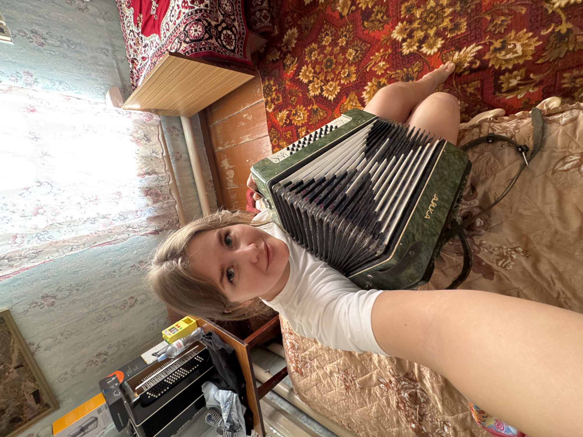 A woman with blond hair wearing a white shirt is holding a green accordion. She is sitting on a bed with a brown patterned bedspread and a red and gold patterned rug is nearby. A wooden table is visible in the background. Another accordion is sitting on a table with a wooden frame, along with a yellow item, a bottle of water, and some clothing. The room has patterned wallpaper. The location may be in Фоки, Russia.