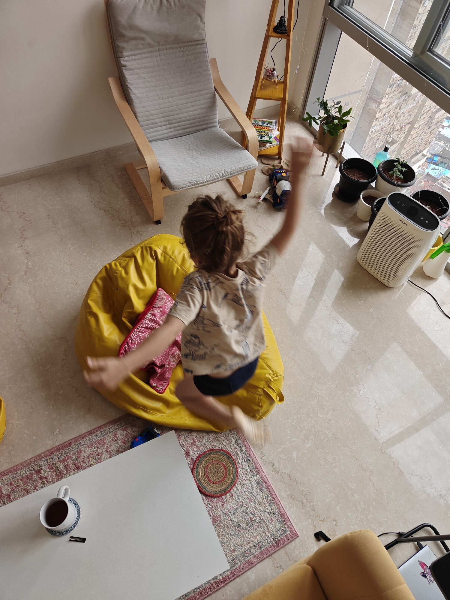 The image depicts an indoor scene, likely an apartment in Mumbai, India, given the urban landscape visible through a large window. In the foreground, a child with brown hair is captured mid-jump, their body blurred with motion, wearing a t-shirt and dark shorts. They are positioned above a large, bright yellow beanbag chair, on which a pink cushion with a floral pattern rests.
To the left of the beanbag, a light-colored armchair with wooden accents and grey upholstery is visible. The floor is tiled in a light, marbled pattern. A patterned rug with red and beige tones lies on the floor, partially beneath the beanbag. On the rug, near the bottom left of the frame, is a white coffee mug filled with a dark liquid, placed on a blue and white patterned coaster, next to a small black object.
In the background, a tall, narrow wooden shelving unit holds various items, including books, a small figurine, and decorative glass. Adjacent to the shelving unit is a large window offering a view of a dense cityscape with tall buildings, characteristic of Mumbai. Several potted plants are placed on the floor near the window, along with a white air purifier unit and a spray bottle. The overall impression is a living space with elements of play, relaxation, and an awareness of the urban environment.