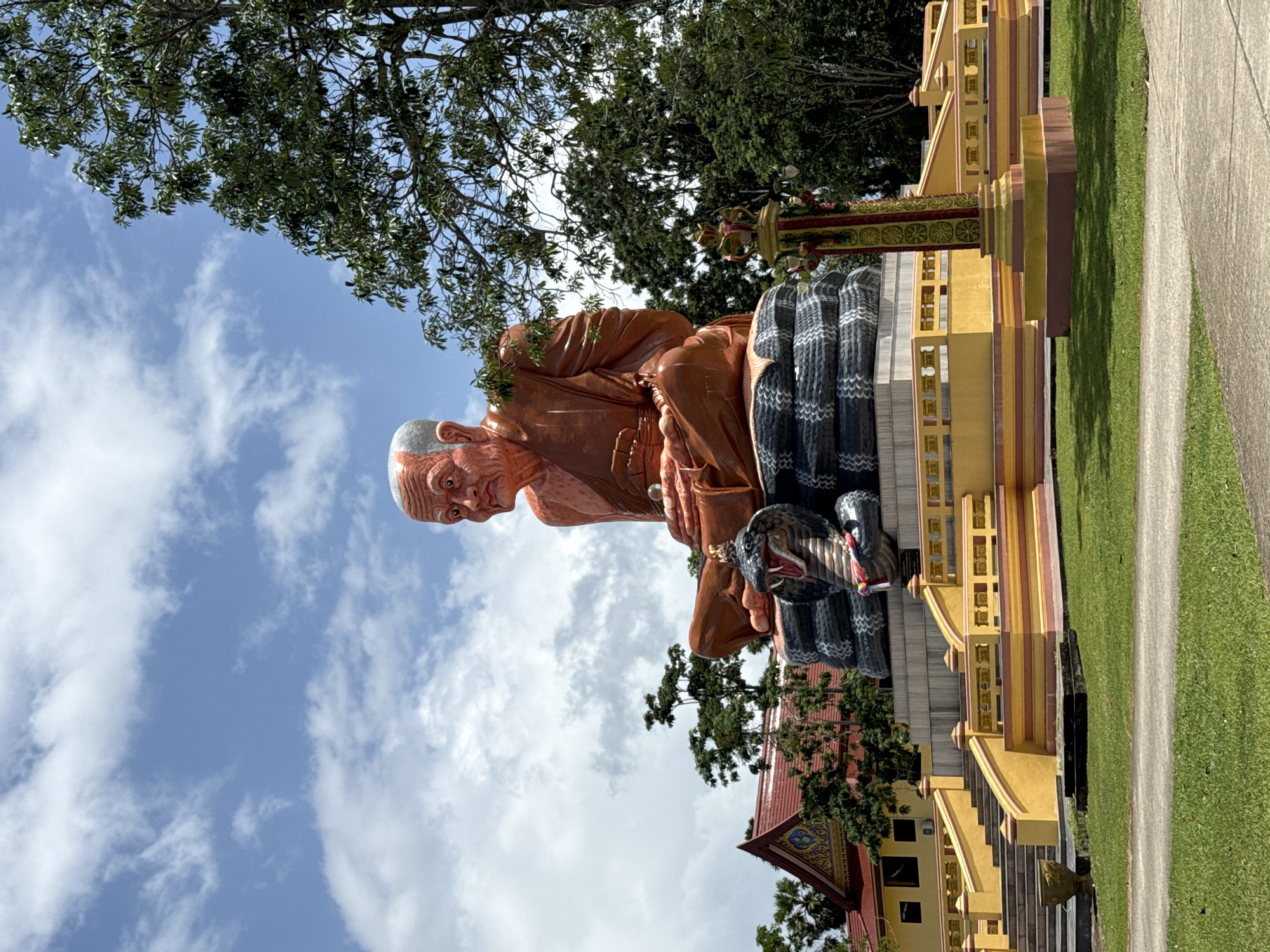 This is a full, eye-level, outdoor, vertical, medium shot of a large, orange-colored statue of a seated Buddhist monk in front of a bright blue sky with scattered white clouds. The monk statue is the dominant subject, positioned centrally and slightly to the left of frame. It is seated in a cross-legged meditative pose on what appears to be a coiled, black-and-white serpent sculpture. The monk's face is wrinkled, and he has a serious expression. He is wearing saffron-colored robes.
To the left of the statue, a yellow and red temple building with a red tiled roof is visible. Behind the statue and to the right, lush green trees with dense foliage are present. In the foreground, a well-maintained green lawn is visible, with a concrete pathway cutting through it. A brightly colored, ornate yellow and red platform with staircases and railings surrounds the base of the statue. A decorative, red and yellow pillar is positioned to the right of the statue's base. The overall setting appears to be a religious or cultural site in an outdoor environment. This statue is located in the city of Na Nuea, Thailand.