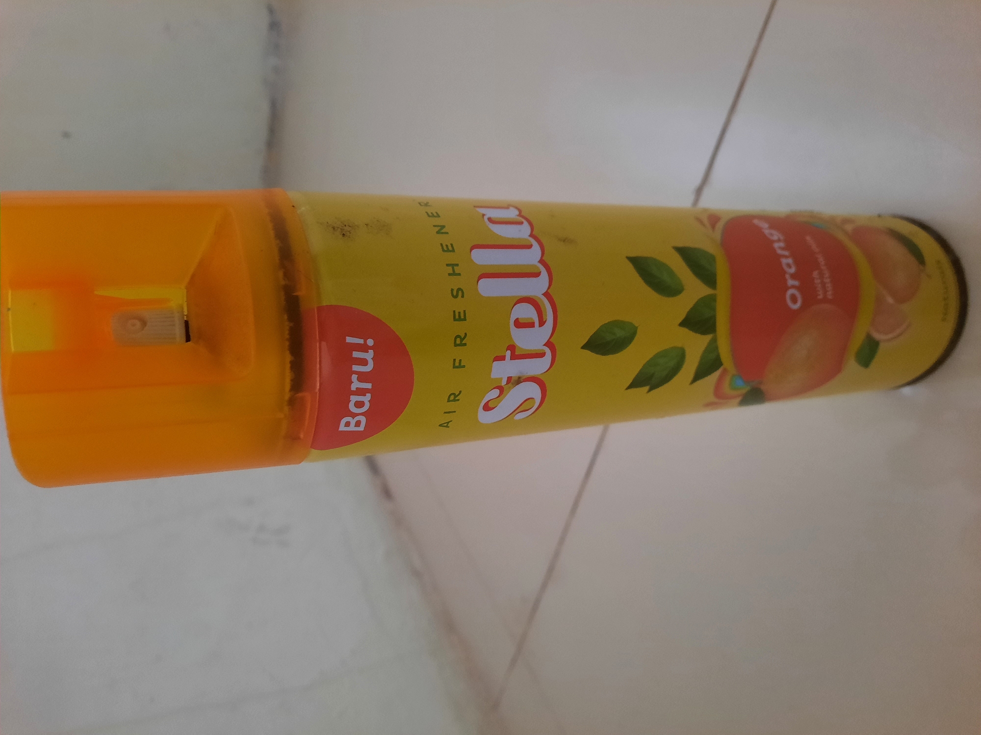 A yellow can of Stella brand air freshener with an orange cap is standing upright on a light-colored tiled floor. The can has the text "Baru!" in a red oval with white lettering. Below that, "AIR FRESHENER" is written in black. The brand name "Stella" is prominent in large white letters with a red outline. Graphics of orange slices and green leaves are depicted on the lower part of the can. The word "Orange" is written in white on a red oval, with smaller text below reading "with natural oils." The background wall is a light, textured color. The scene is likely indoors, in a room in Lhokseumawe, Indonesia, given the tiled flooring typical of Indonesian households or public spaces.