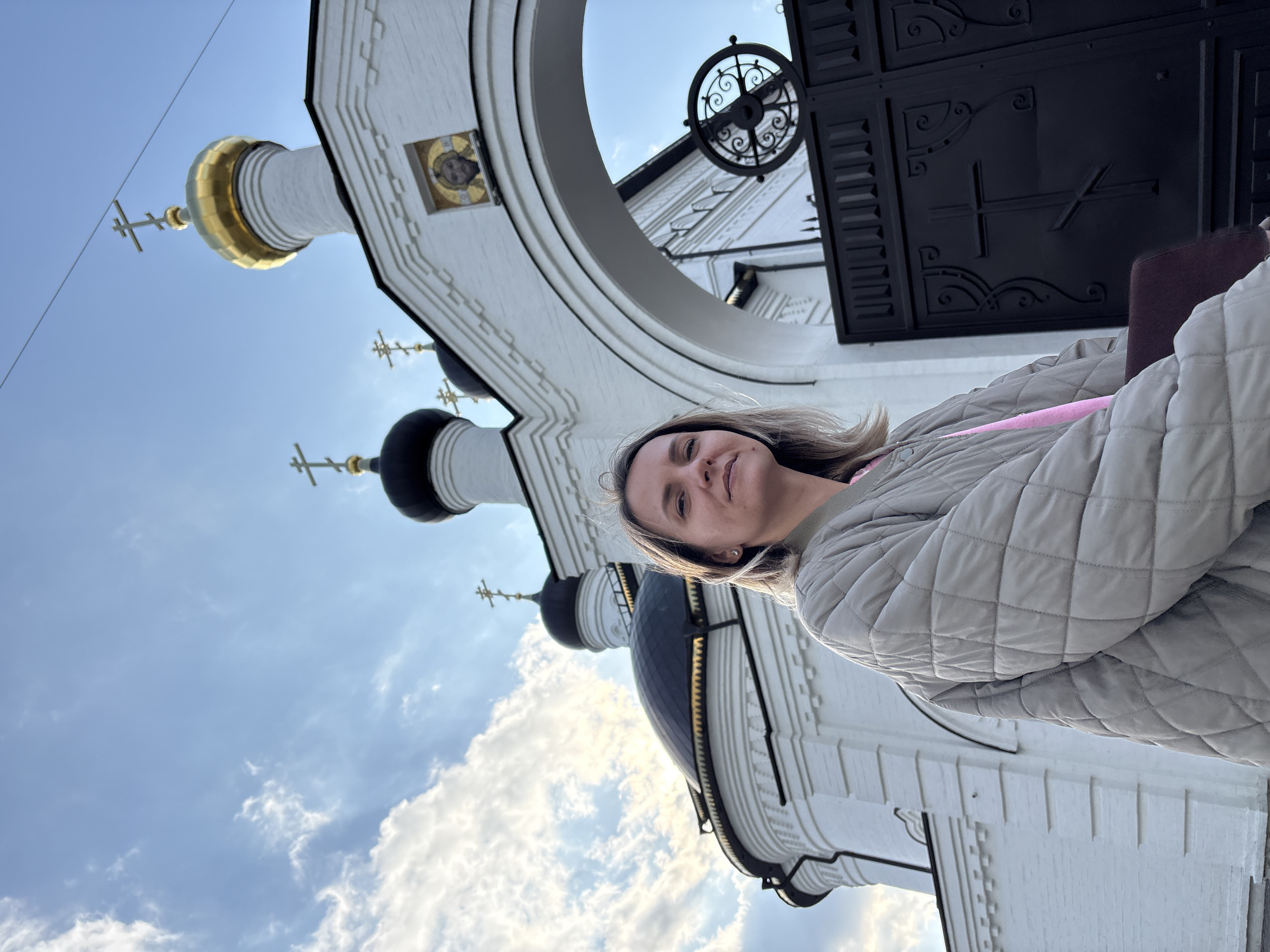 A woman stands in front of the Holy Resurrection-Nefedovsky Monastery in Ulyanovsk, Russia. The monastery features white walls, black domed roofs, and golden crosses. The sky above is blue with scattered white clouds. The woman wears a grey quilted jacket with a pink strap showing. A wrought-iron gate with a large cross design is visible above her.