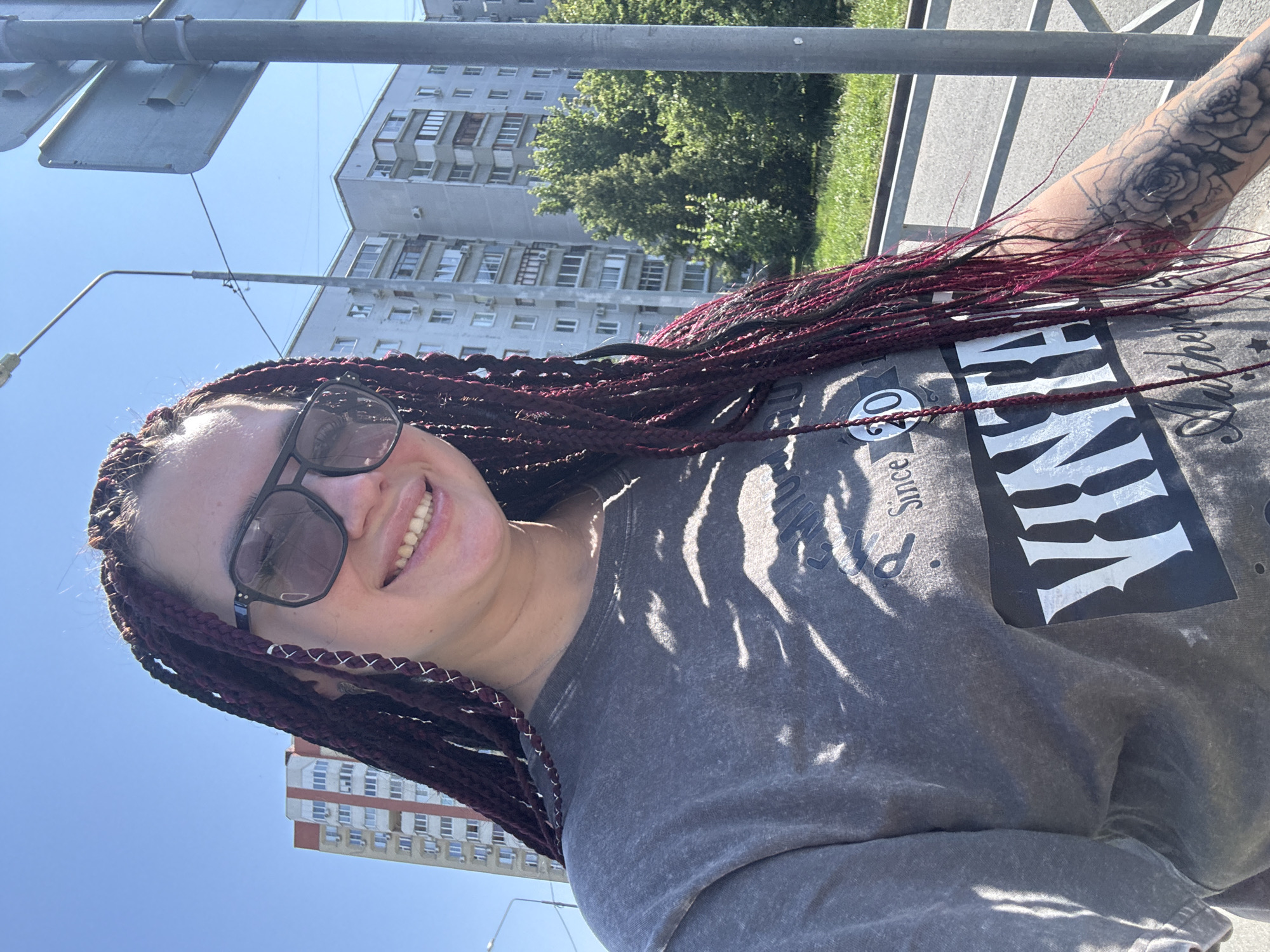A young woman with dark braided hair is taking a selfie outdoors on a sunny day. She is wearing sunglasses and a grey t-shirt with a graphic print. Behind her, there are apartment buildings and trees. The scene appears to be a street or sidewalk in a residential area of Tolyatti, Russia. A metal railing and a lamppost are also visible in the background.
