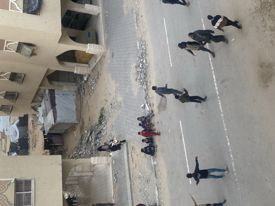 An aerial view shows a group of people walking or standing on a road next to a building. Some people are clustered together, while others are more spread out. Debris and rubble are visible on the side of the road, near the building. The building appears to be multi-story with distinct architectural features, suggesting a residential or commercial structure. Laundry hangs outside one part of the building, possibly indicating displacement.