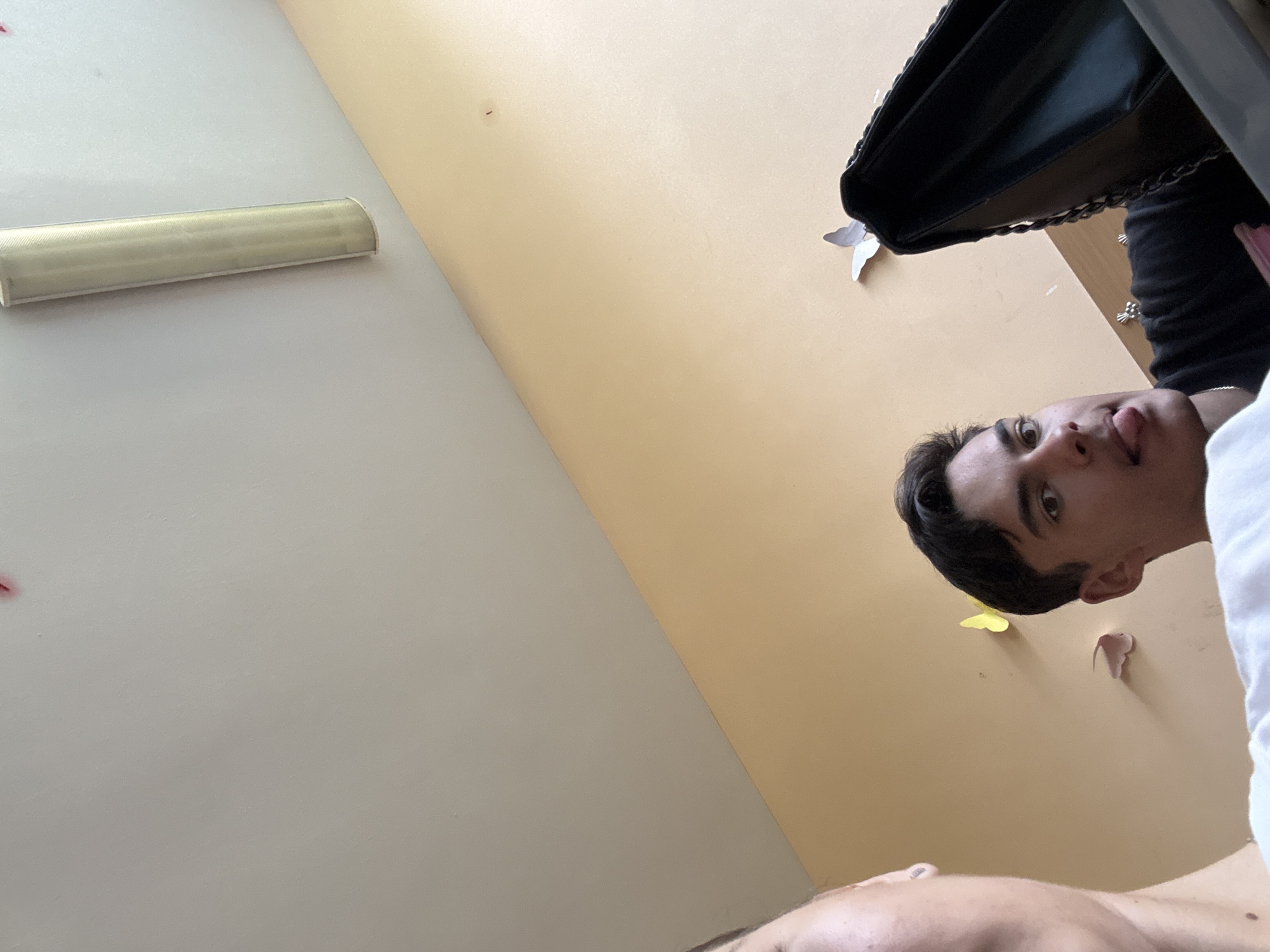 The image, taken from a low angle looking up, shows two people and a black purse. One person is in the right side of the frame, their face clearly visible with their tongue sticking out. The other person is mostly out of frame, with just the top of their head and part of their torso visible at the bottom. A black purse is visible in the upper right corner of the image. The background includes a white wall with a fluorescent light fixture and a beige wall adorned with decorative butterfly cutouts. The location is likely indoors. There is insufficient information to determine the location as Sofia, Bulgaria.