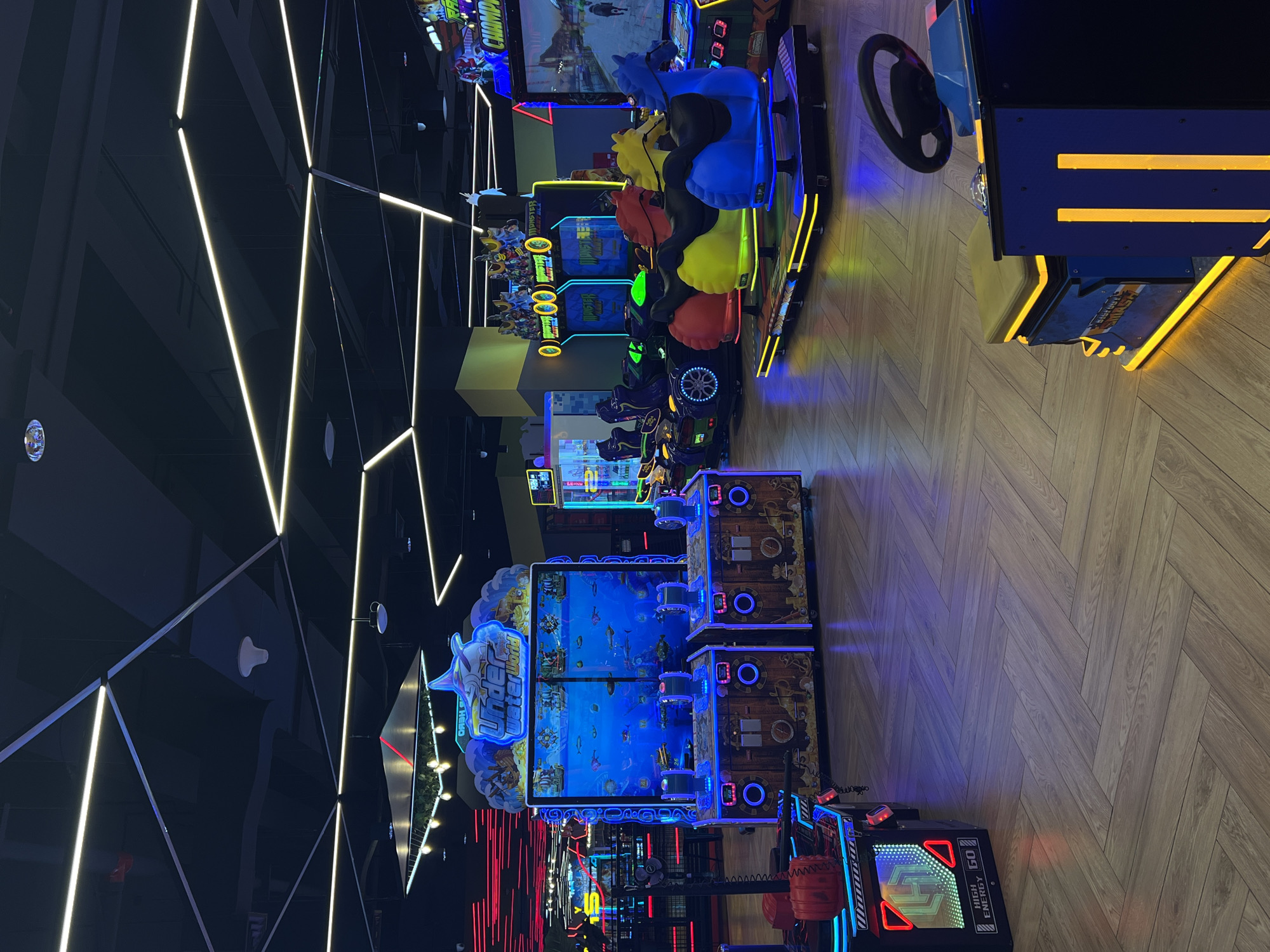 The image depicts the interior of an amusement arcade in Kuta, Indonesia. The scene is dominated by various arcade game machines, characterized by their bright, colorful lights and electronic displays. Prominently featured are two large "Under Water World" shooting gallery games, each with a screen displaying an underwater scene and two player stations with controls. To the right, there are several kiddie rides shaped like horses and cars, illuminated with neon lights. In the background, more arcade games and seating areas are visible, along with a distinctive ceiling structure with geometric lighting. The flooring is a herringbone pattern wood-look laminate. There are no people visible in the frame.