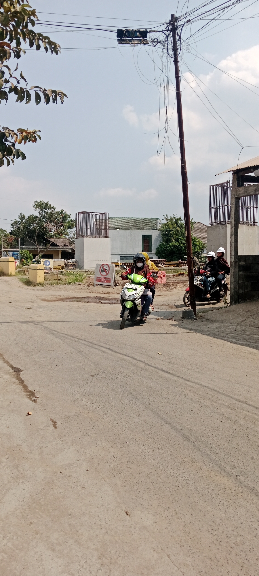 In Purwomartani, Indonesia, on a bright, clear day, two motorcycles are traveling on an asphalt road. The first motorcycle, a green and white scooter, carries two individuals, one driving and one as a passenger. Both are wearing helmets. The driver wears a brown jacket and a black helmet, while the passenger wears a yellow headscarf and a dark helmet. They are approaching a traffic sign that depicts a car with a red circle and a line through it, indicating "No Cars Allowed." The second motorcycle, a red scooter, is further down the road and also carries two people, both wearing white helmets. The environment is a semi-urban area with a construction site visible in the background. The construction site features large concrete pillars with rebar structures, suggesting an ongoing project. There are trees and some low-lying vegetation. A utility pole with a complex network of wires stands to the right of the frame, with a transformer box attached to it. The road surface shows signs of wear and tear, with some scattered debris.