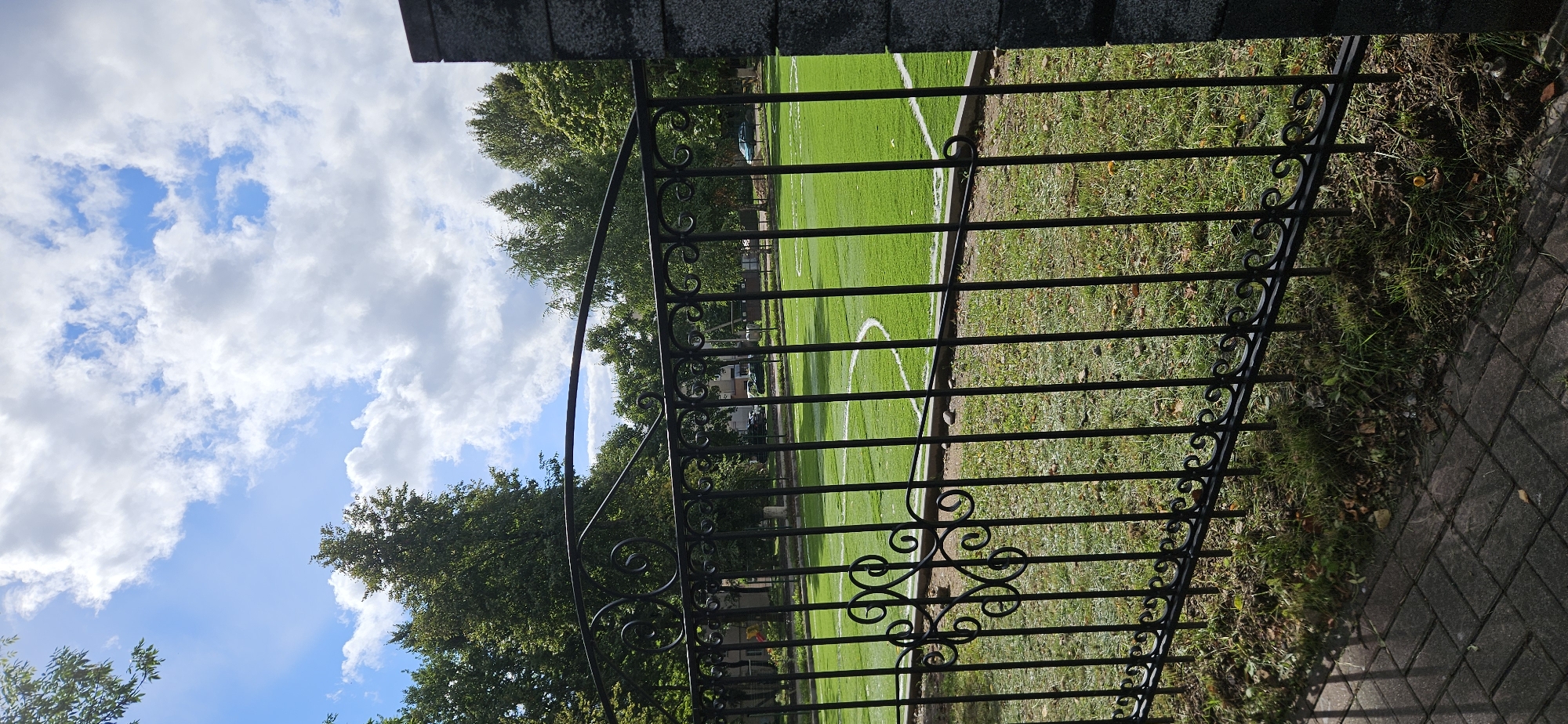 An outdoor, vertical, eye-level shot captures a view through a black wrought-iron fence toward a bright green, artificial turf sports field. The field is marked with white lines, suggesting it is a soccer or football pitch. Beyond the field, a line of mature deciduous trees with full, green foliage forms a backdrop. The sky above is a vibrant blue, dotted with fluffy white cumulus clouds. The foreground features a paved walkway made of grey interlocking bricks, with patches of grass and scattered leaves along the base of the fence. A dark, textured concrete pillar marks the right edge of the frame. The overall impression is one of a well-maintained urban park or recreational area on a bright, partly cloudy day.